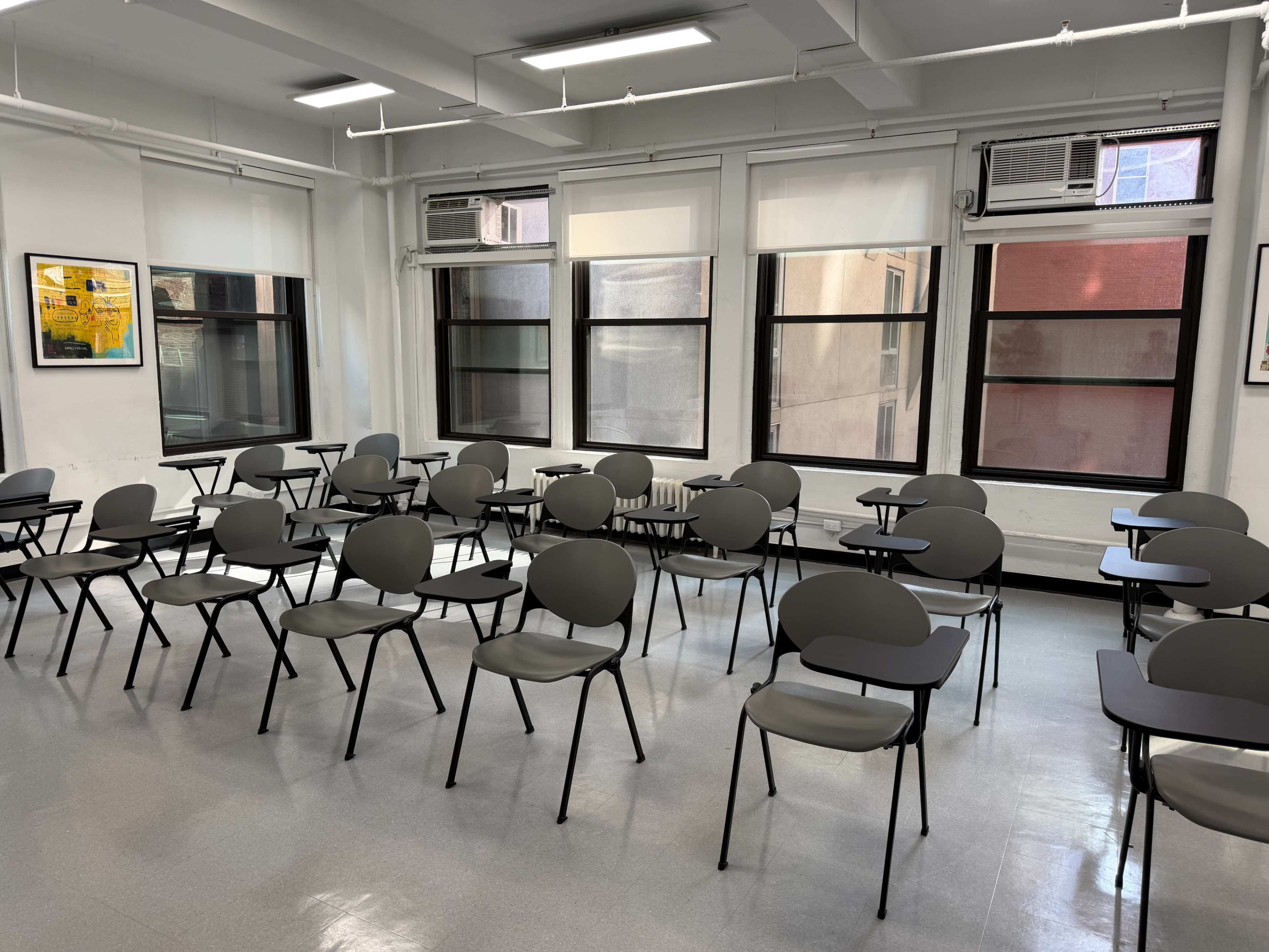 The image depicts a classroom filled with neatly arranged chairs and desks, facing windows that provide natural light.