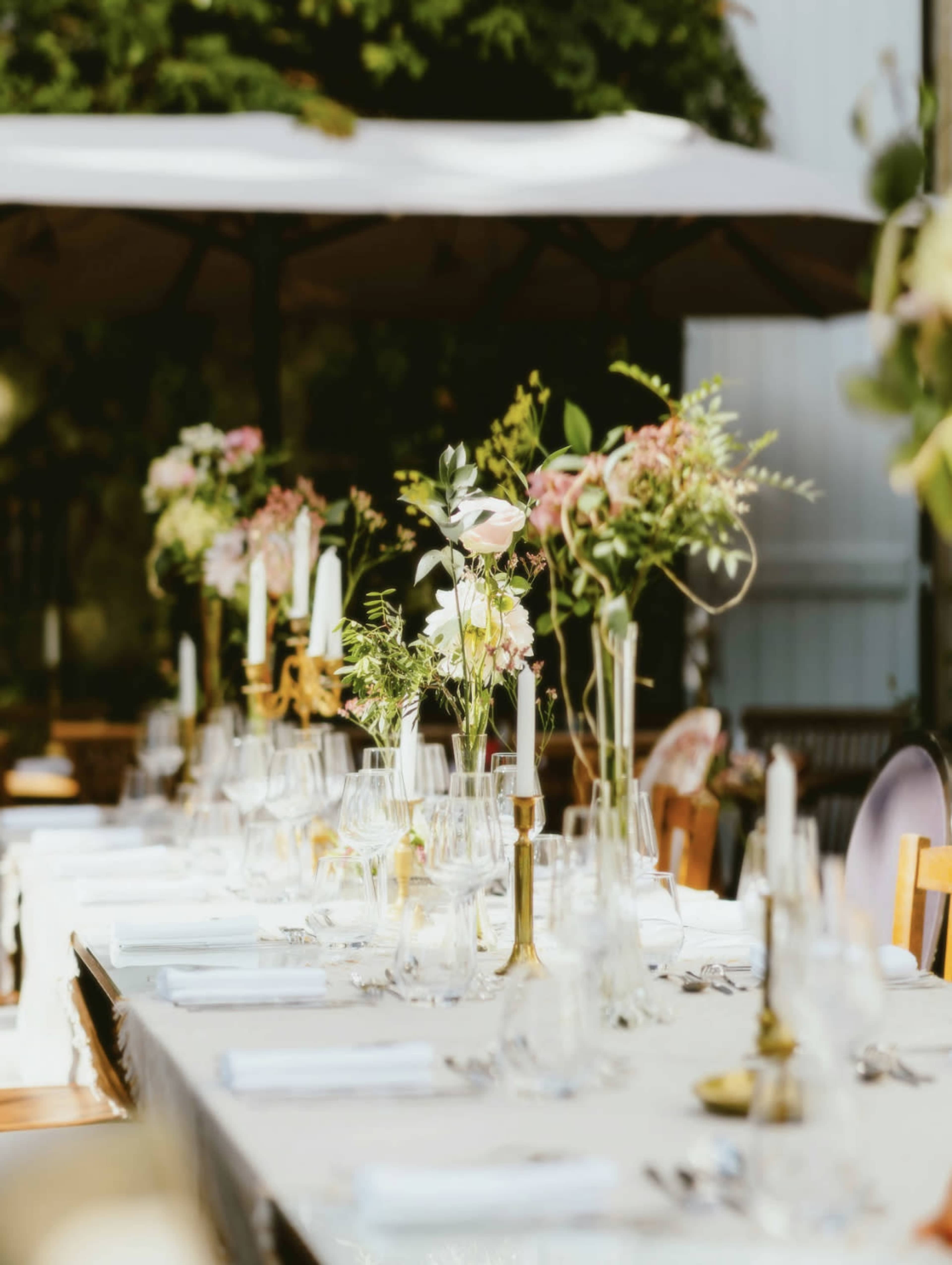 A long dining table is beautifully set outdoors, adorned with elegant floral arrangements and candles under a canopy of umbrellas.
