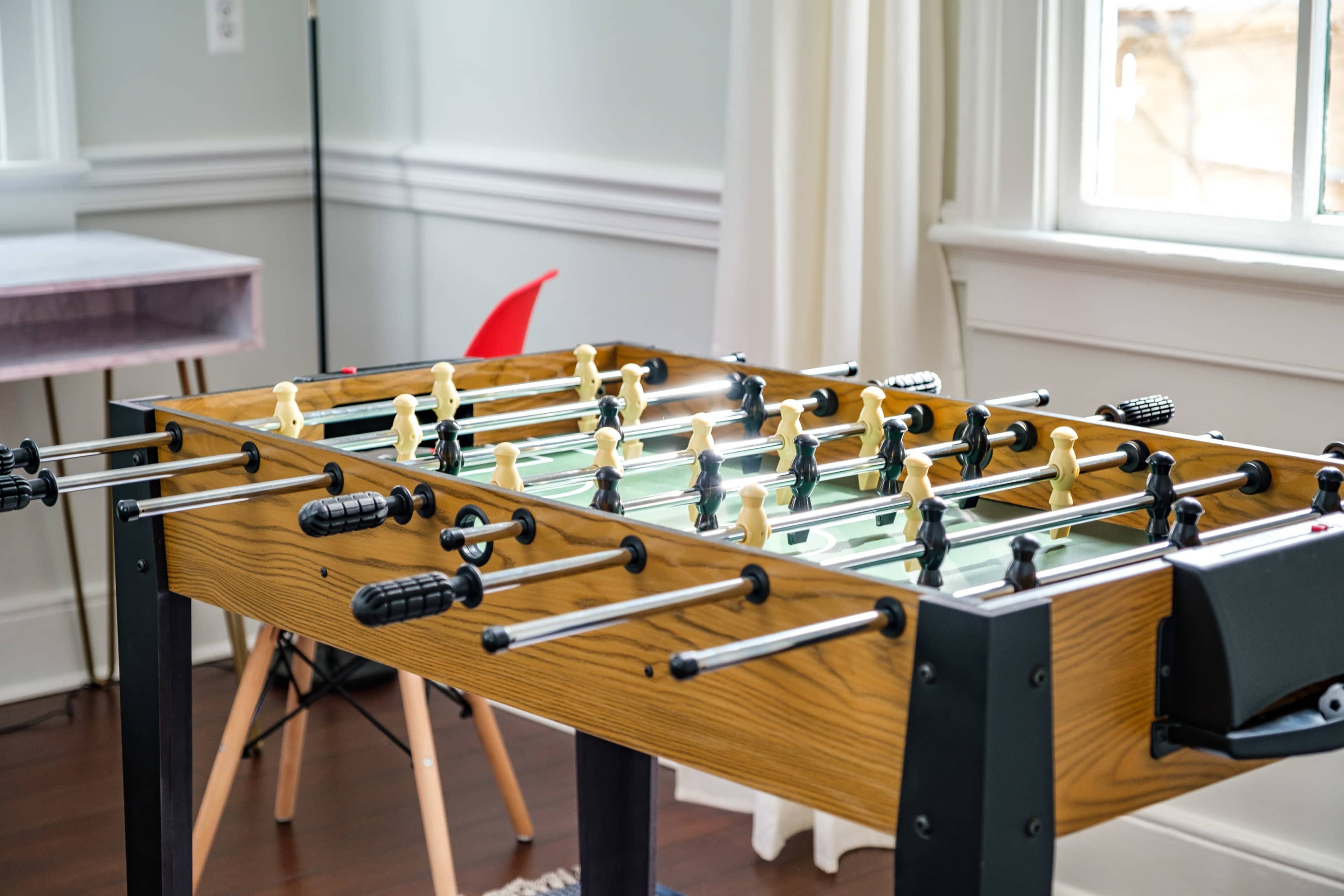A foosball table is positioned in a well-lit room with wooden flooring and a large window.