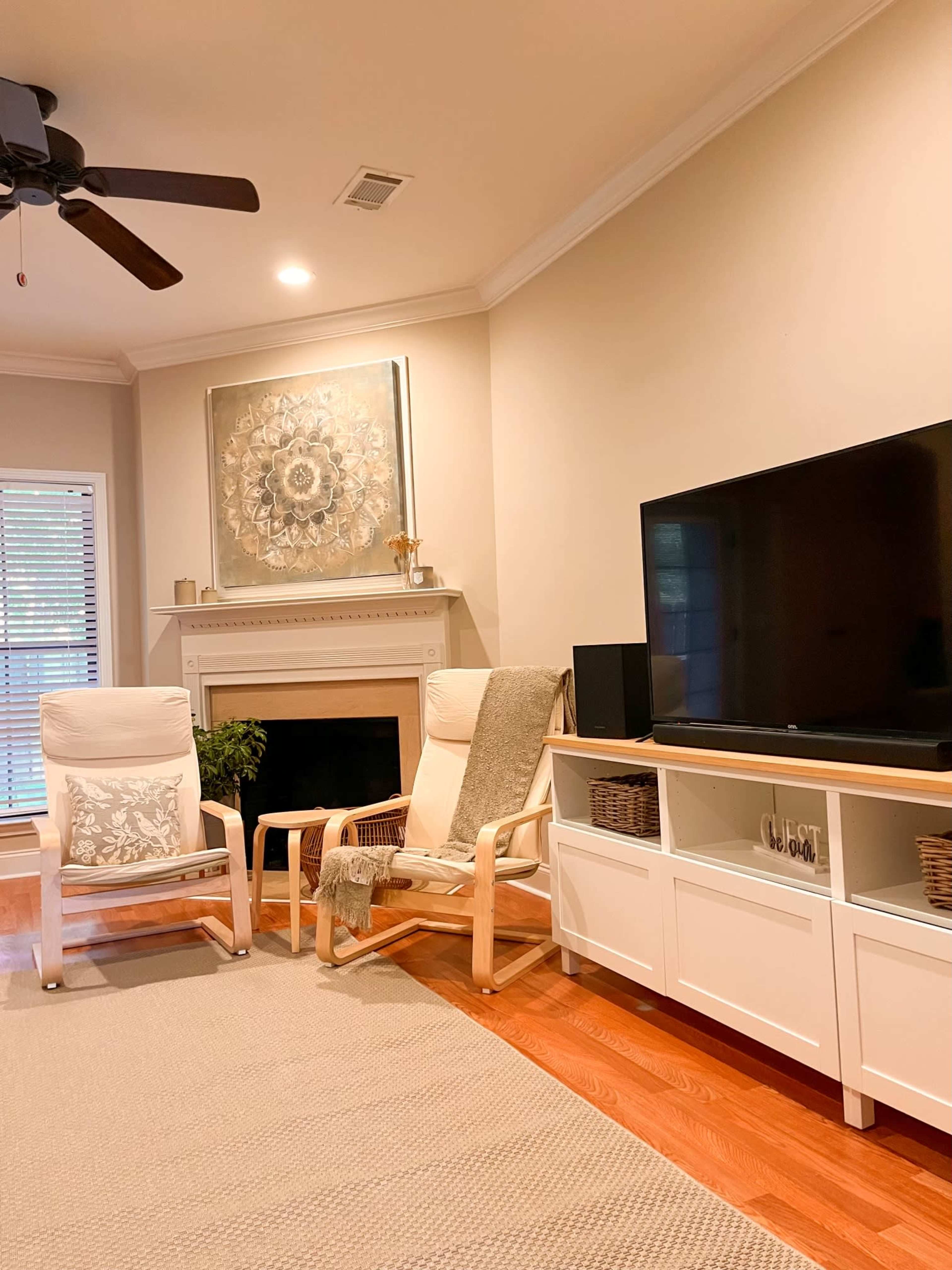 A living room features two wooden chairs, a television on a white cabinet, and a fireplace with a decorative piece above it.
