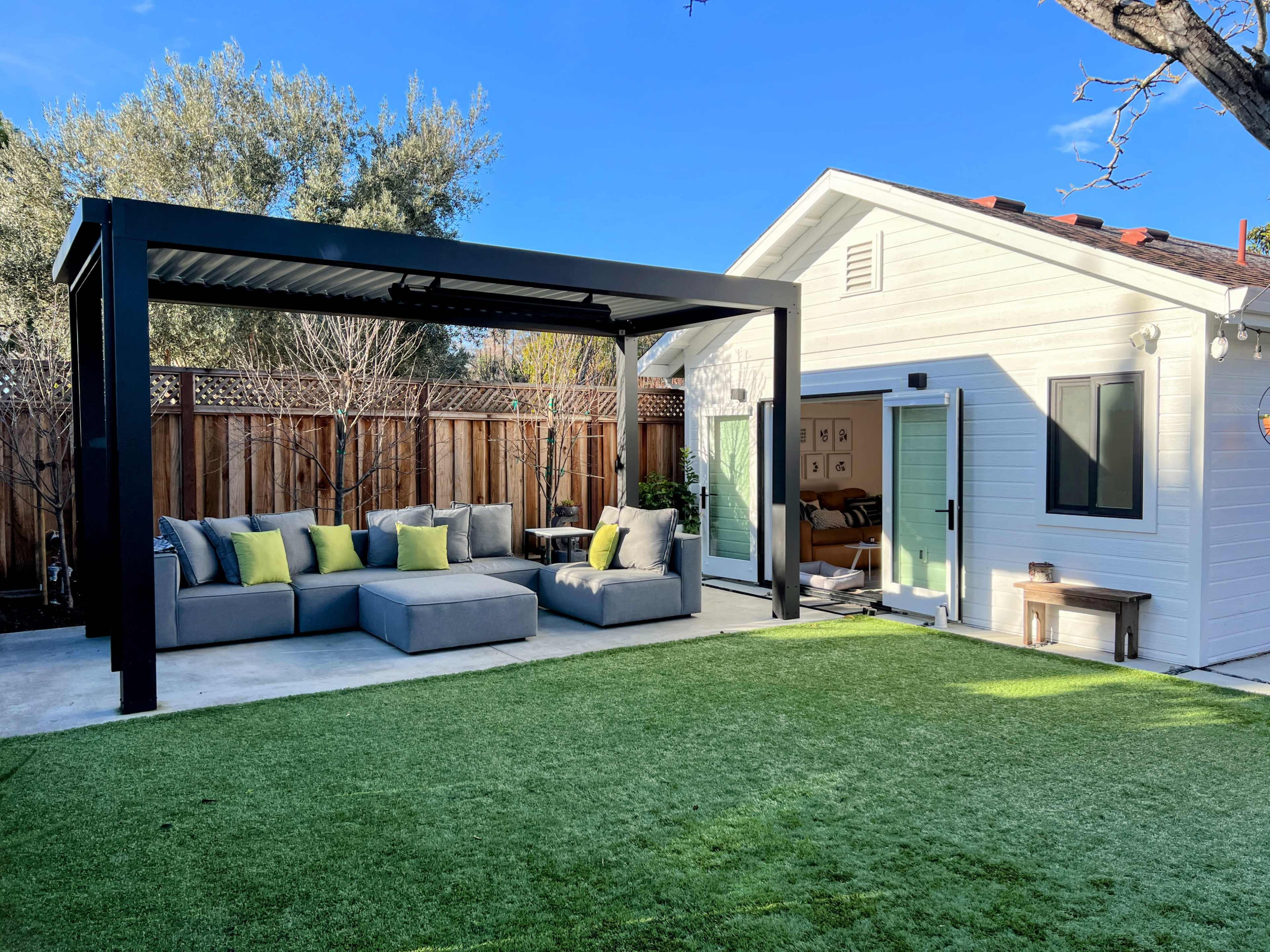 A modern outdoor seating area with a shaded lounge set and a white house with a sliding glass door, surrounded by green grass and trees.
