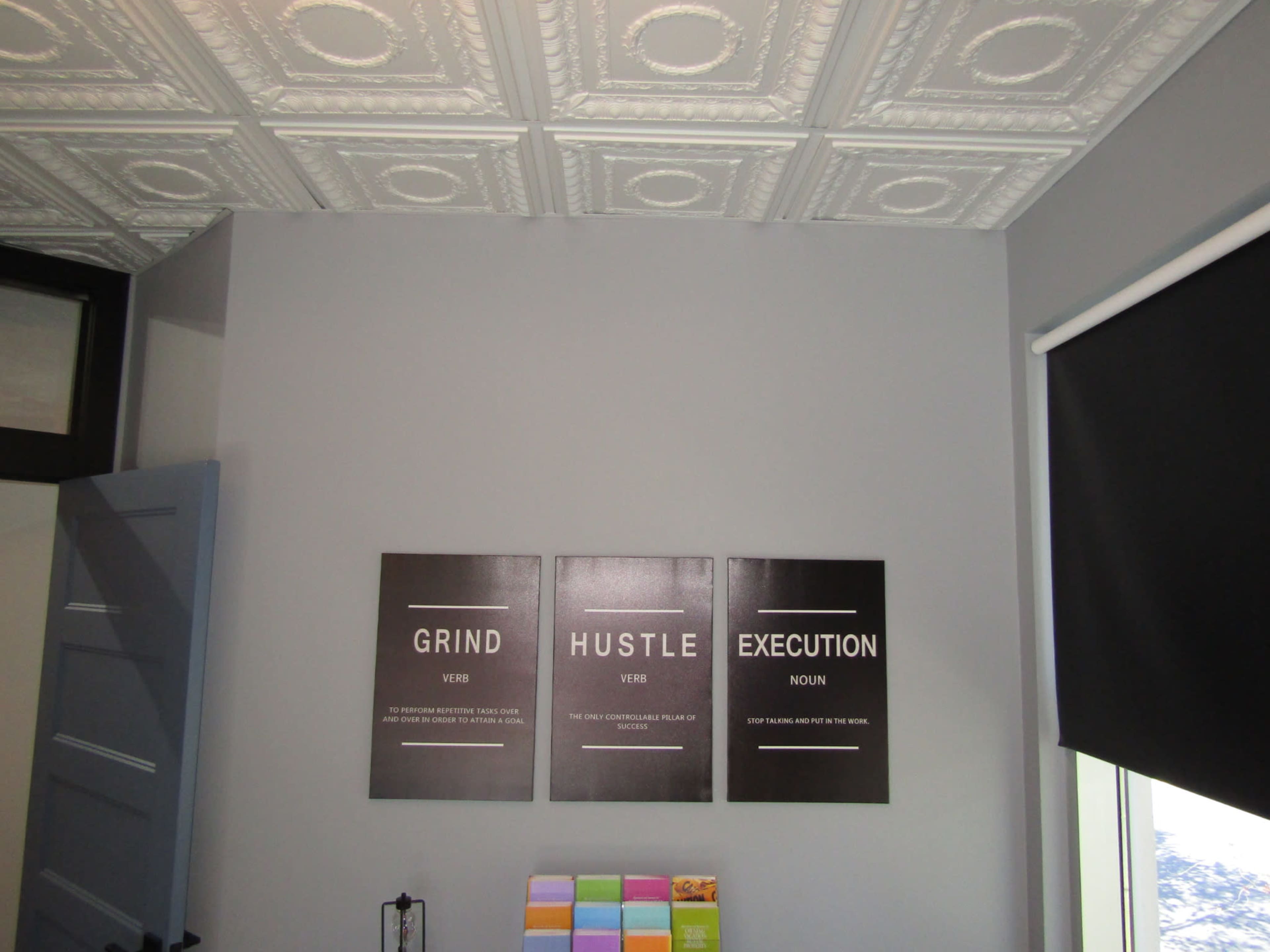 A gray wall displaying three framed signs with the words "GRIND", "HUSTLE", and "EXECUTION" along with their definitions, next to a colorful stack of paper.