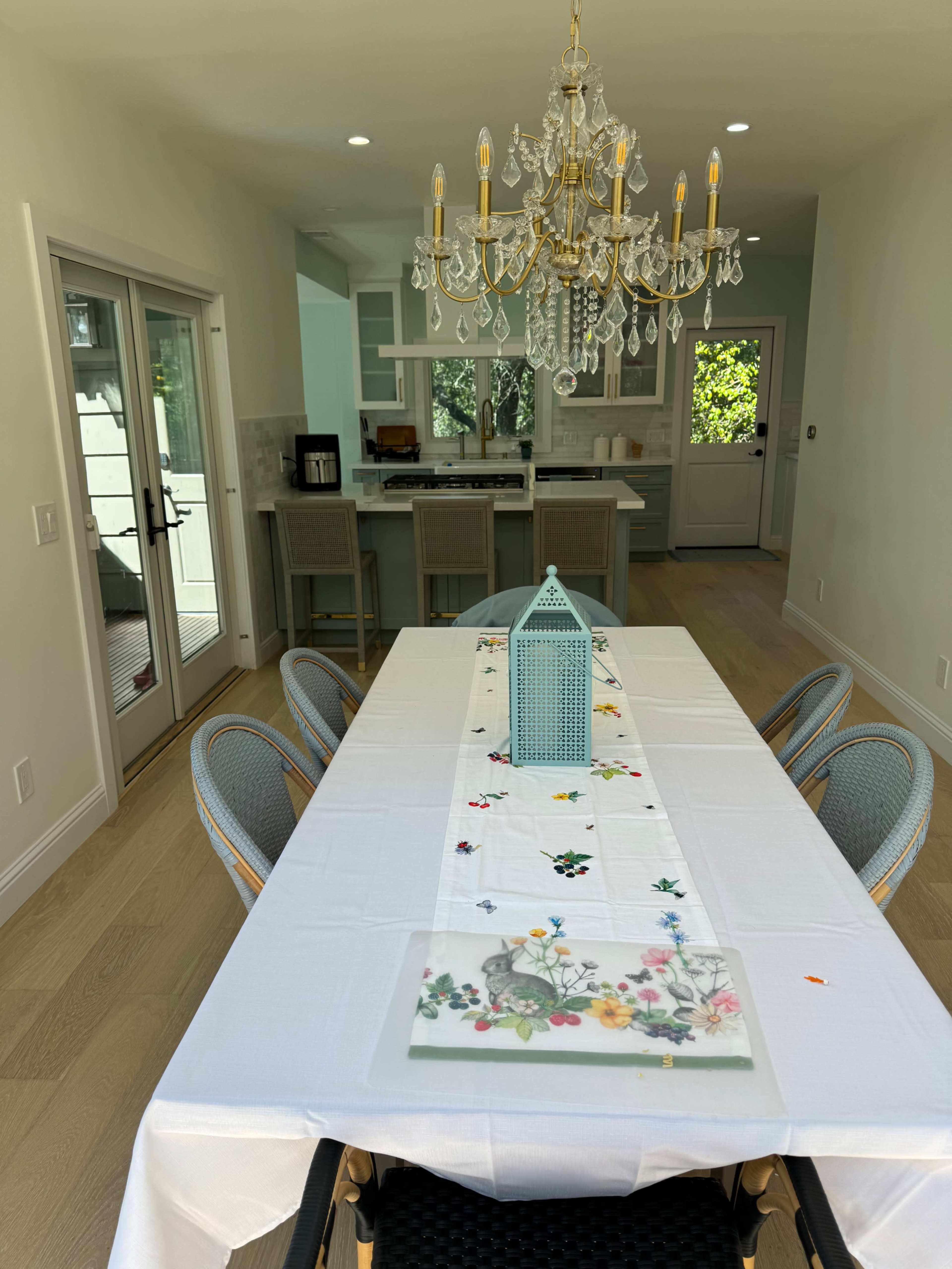 A long dining table set with a floral table runner is illuminated by a chandelier, while a kitchen area is visible in the background.