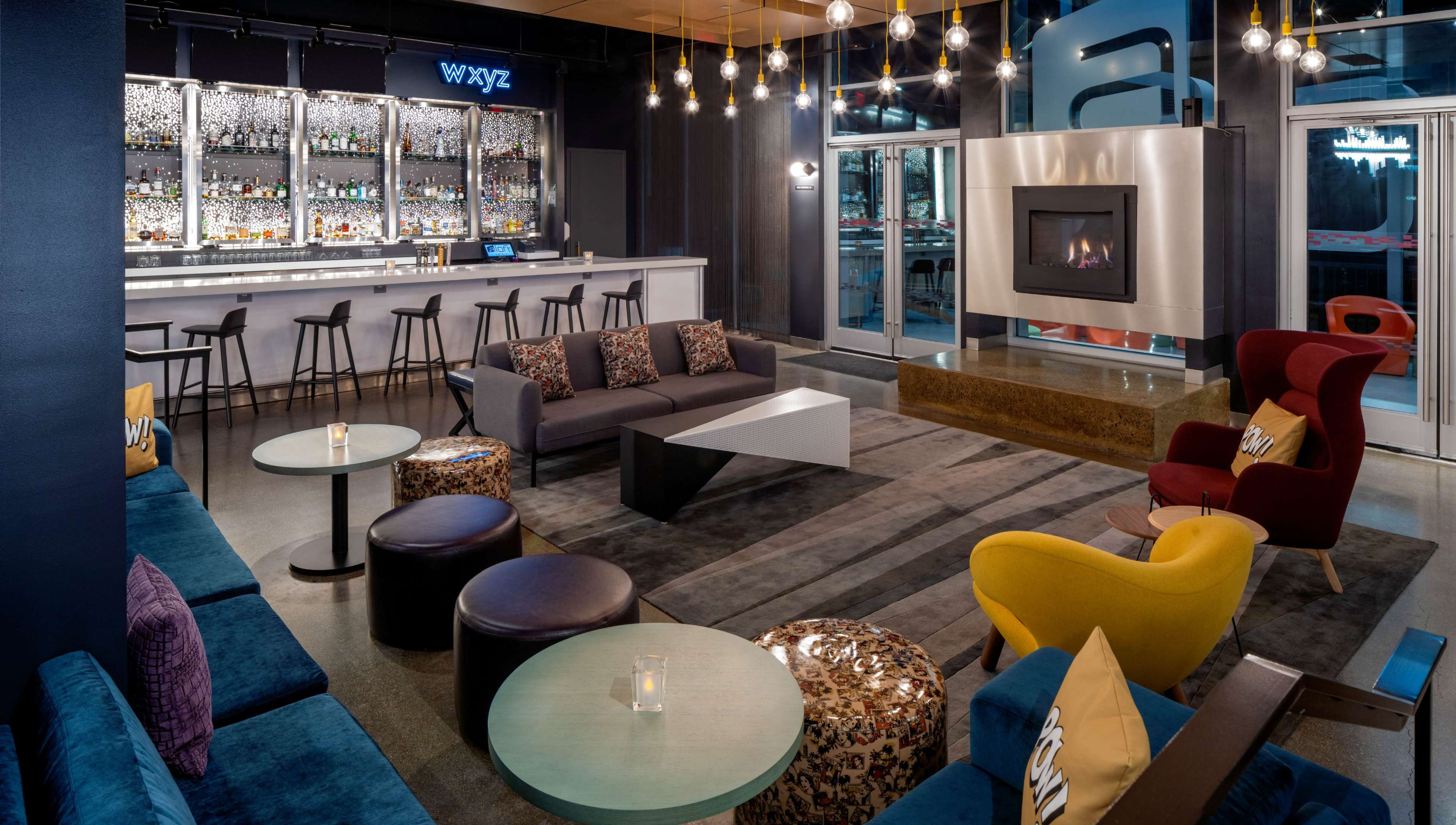 A modern bar area featuring seating options, a fireplace, and a well-stocked backlit bar with various bottles.