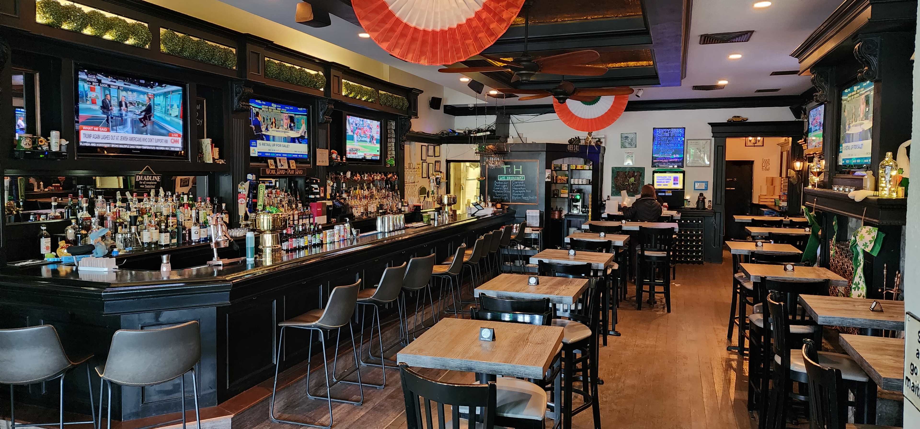 A spacious bar area with a long black counter, several high chairs, and multiple televisions mounted on the walls displaying sports events, while patriotic decorations hang above.