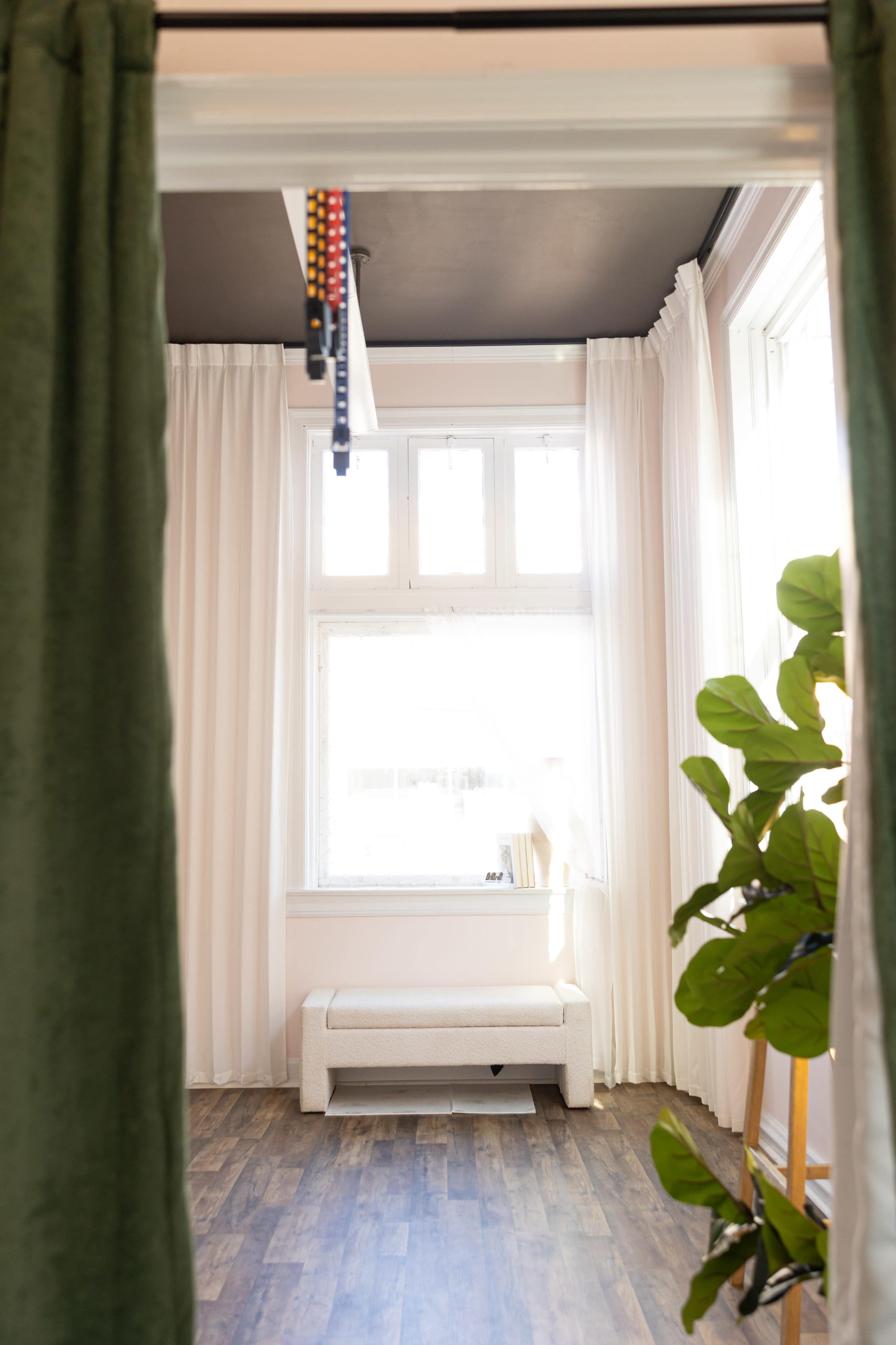 The image shows a room with white curtains, a bench near a window, and a potted plant in one corner.