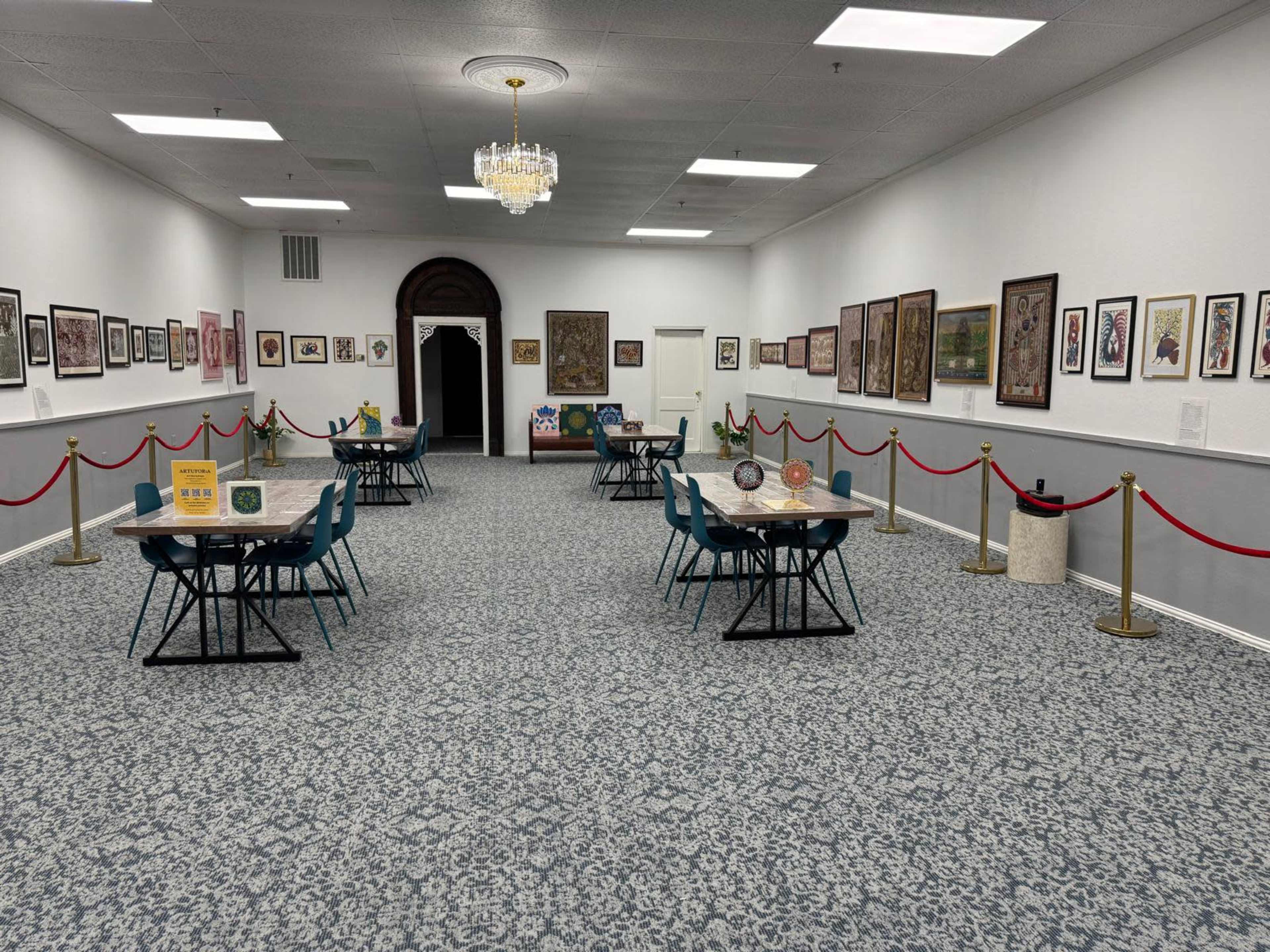The image shows a spacious art gallery with turquoise chairs and tables arranged in a symmetrical layout, surrounded by framed artworks on the walls and red rope barriers defining the walking area.