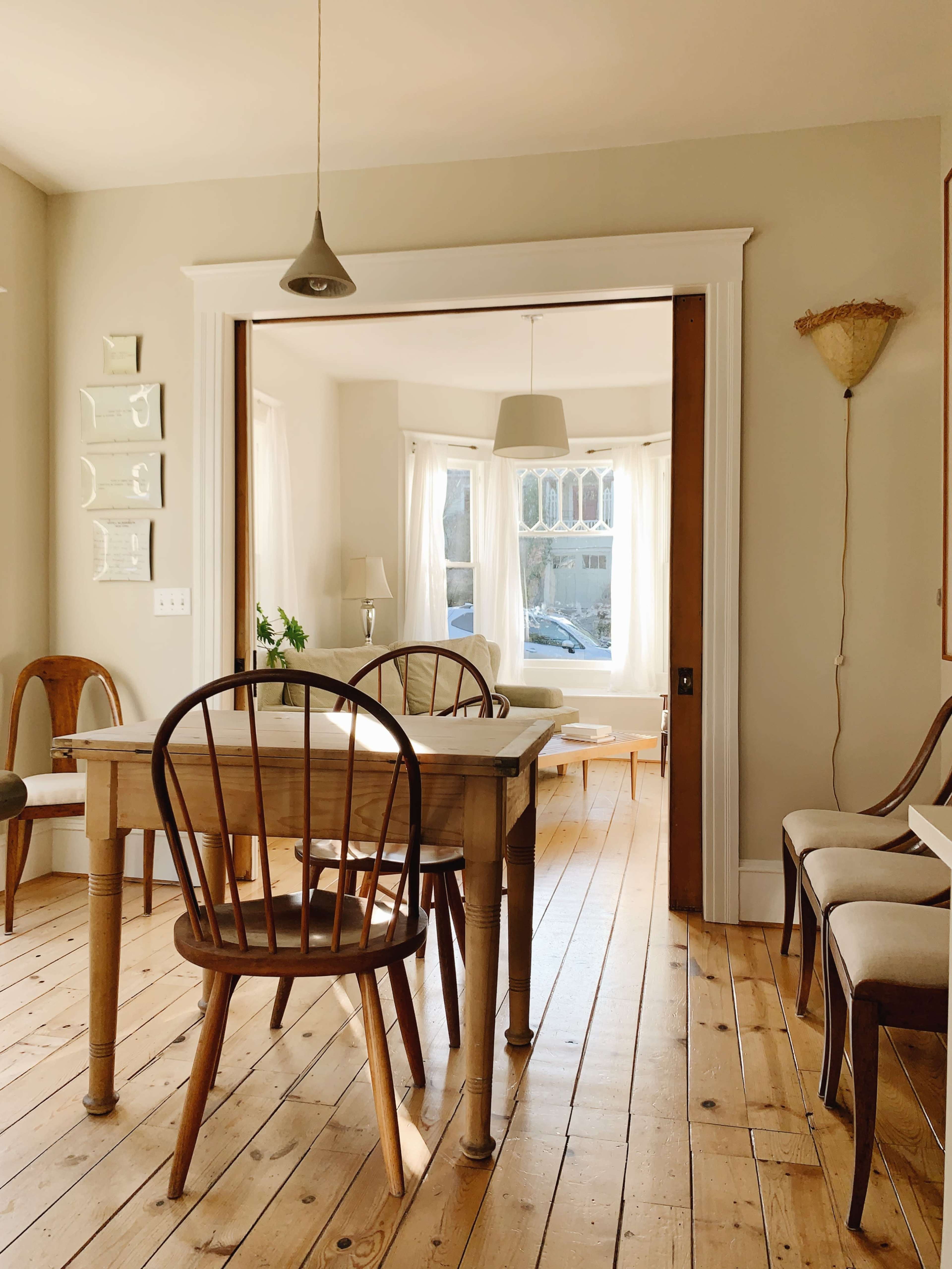 A bright dining area features a wooden table surrounded by chairs, with a view into a sunlit room through an open doorway.