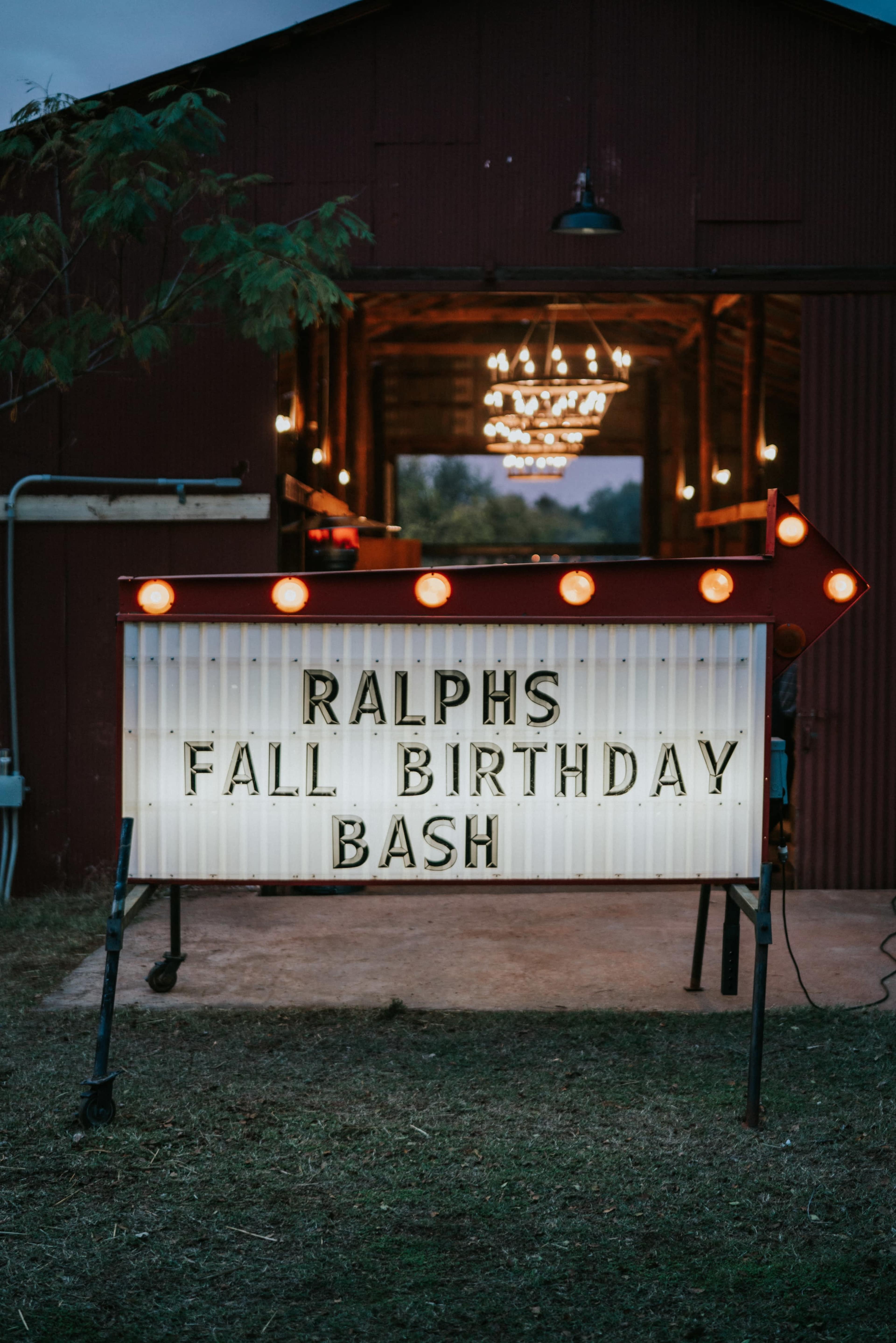 A sign announcing "RALPH'S FALL BIRTHDAY BASH" stands outside a barn lit with hanging lights.