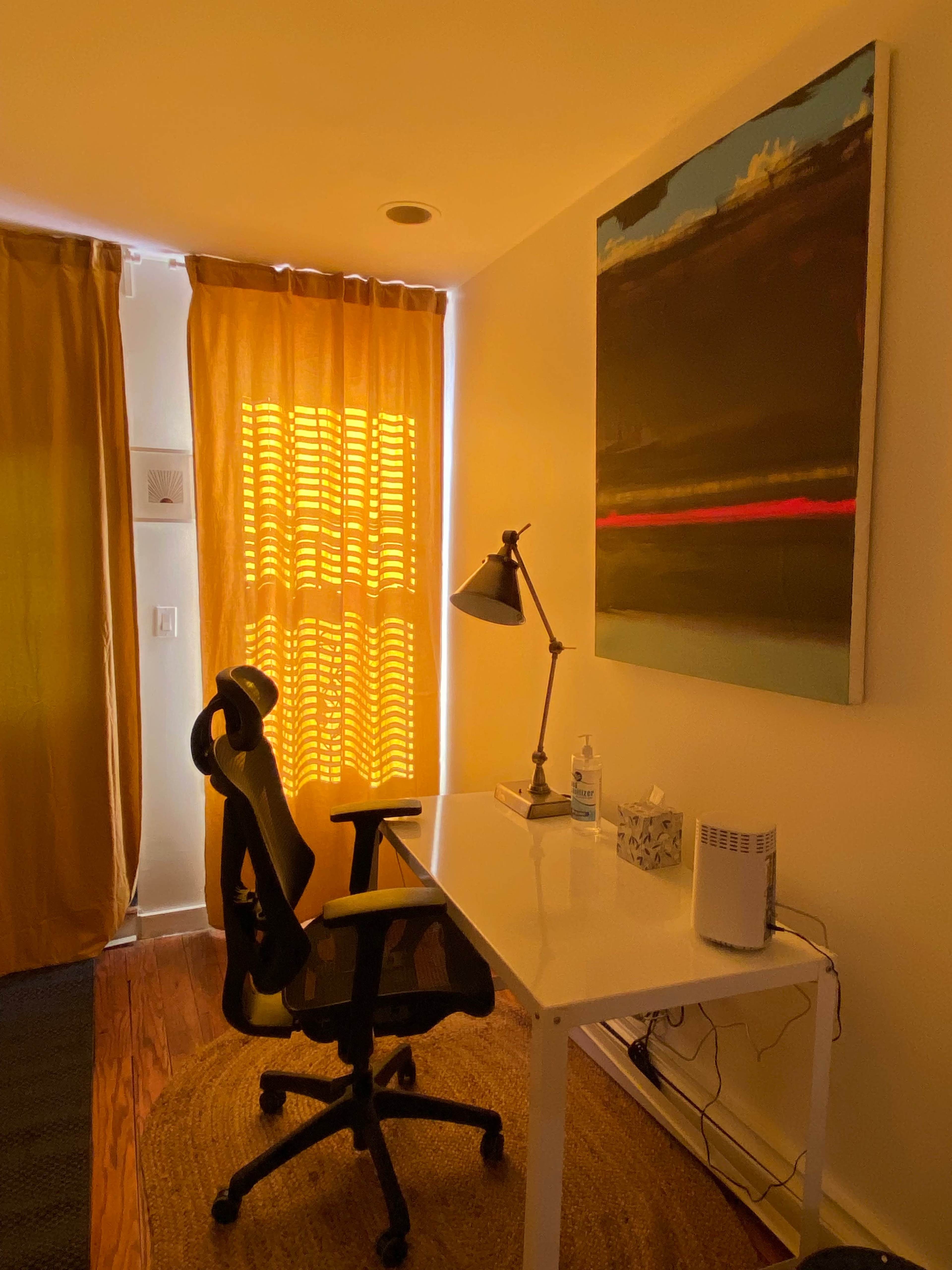A simple workspace features a white desk with a black chair, a lamp, and a painting on the wall, illuminated by light filtering through yellow curtains.