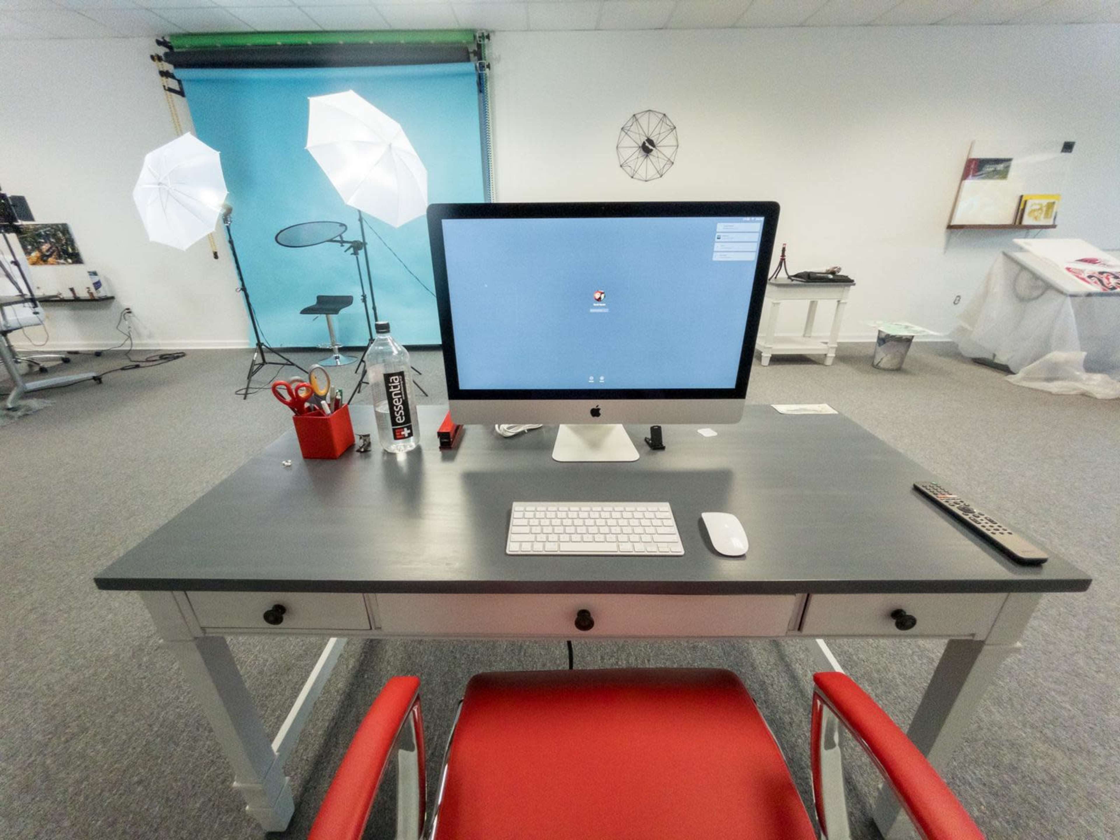 A desk with an iMac computer, keyboard, and various accessories is positioned in a bright studio space featuring photography lighting setups in the background.
