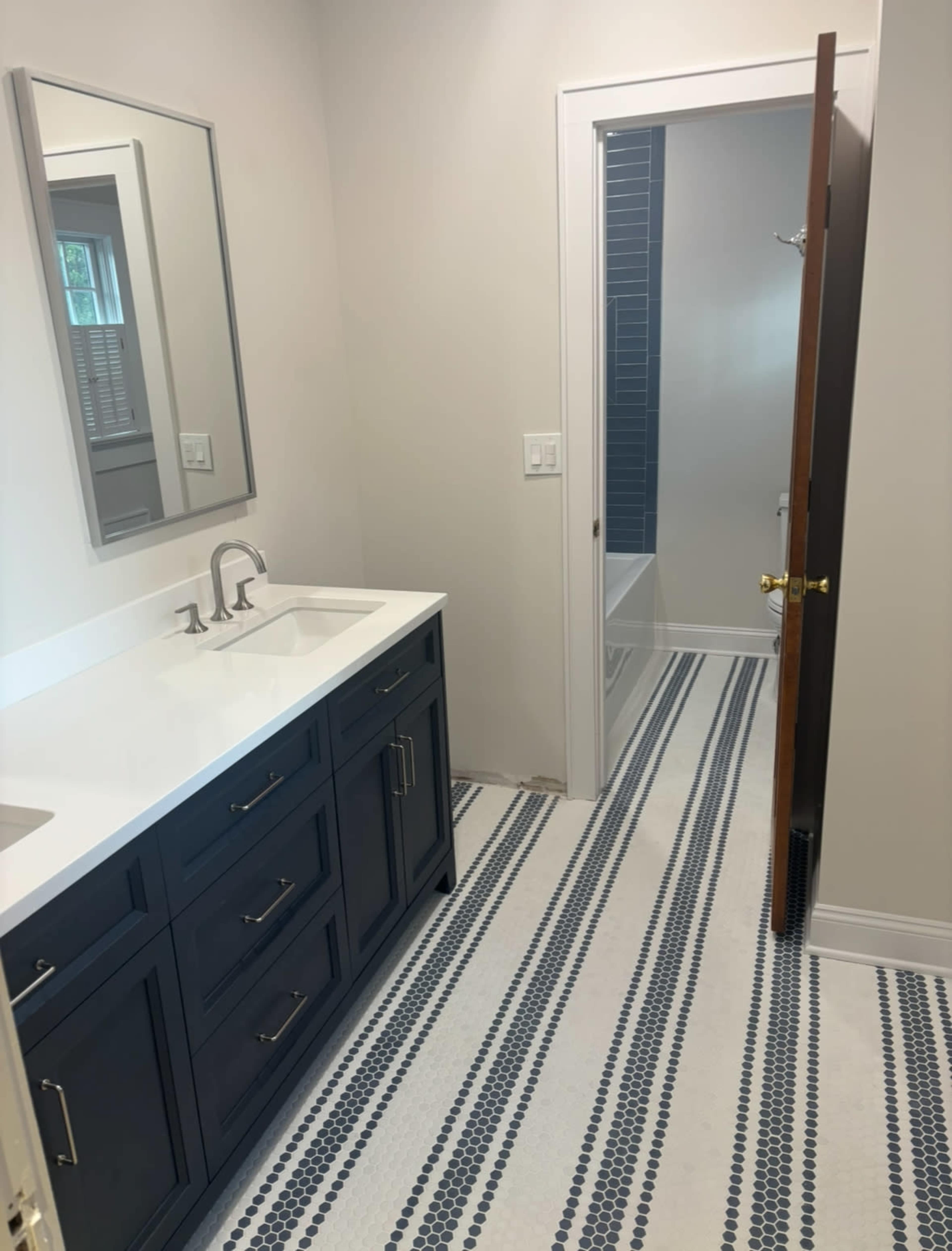The image shows a modern bathroom with a dark blue double vanity, a large mirror, and a doorway leading to a tiled shower area.