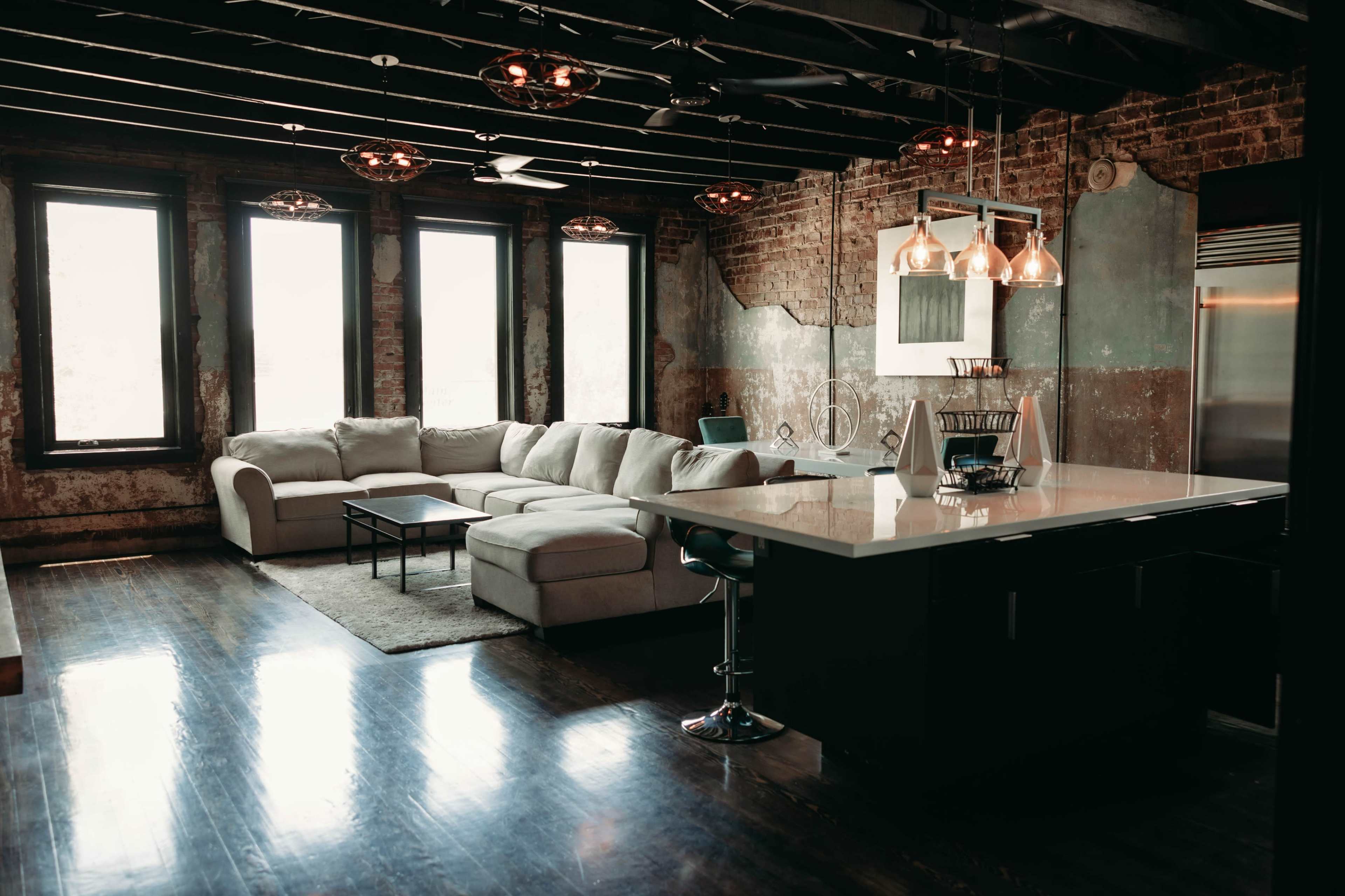 A spacious living room features a large sectional sofa, a coffee table, and a kitchen island with pendant lighting, all set against exposed brick walls and large windows.