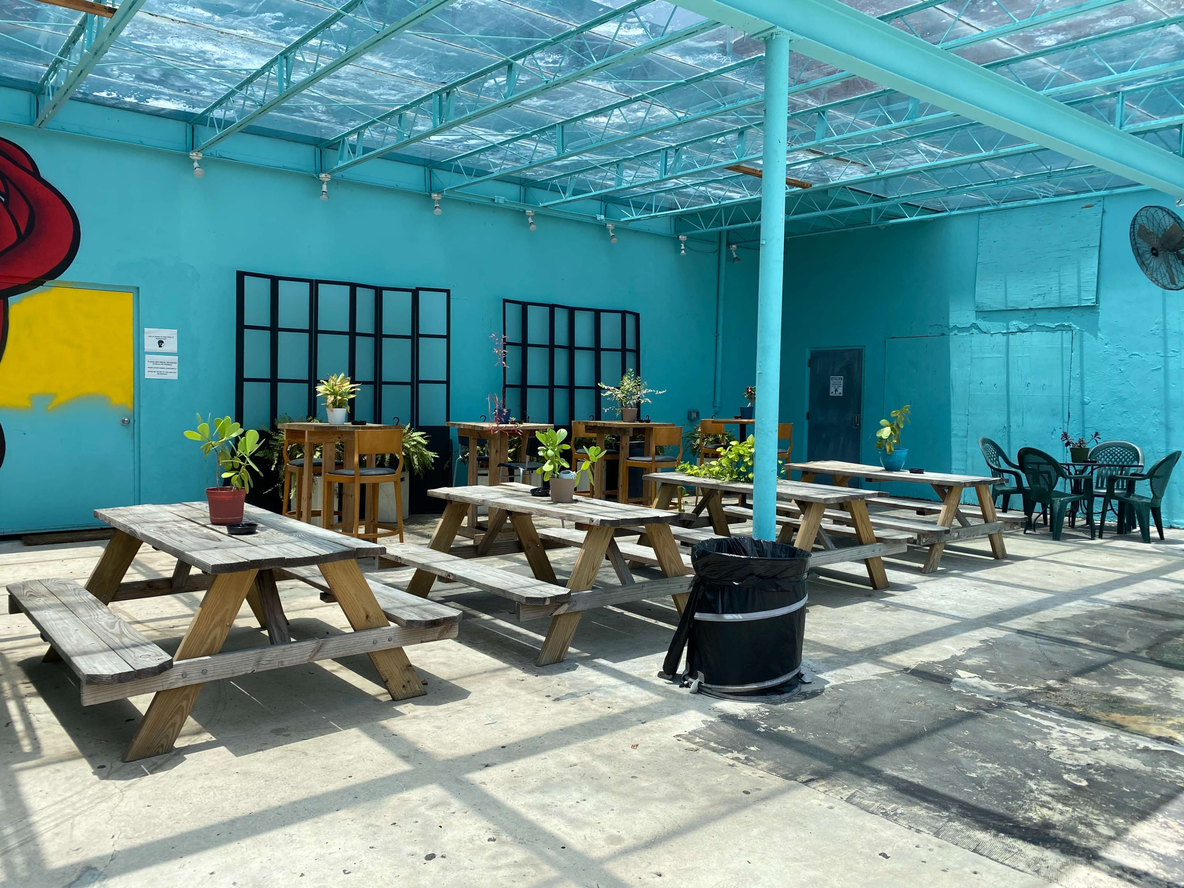 The image shows a vibrant, open seating area featuring wooden picnic tables and potted plants under a transparent roof, with a turquoise wall in the background.