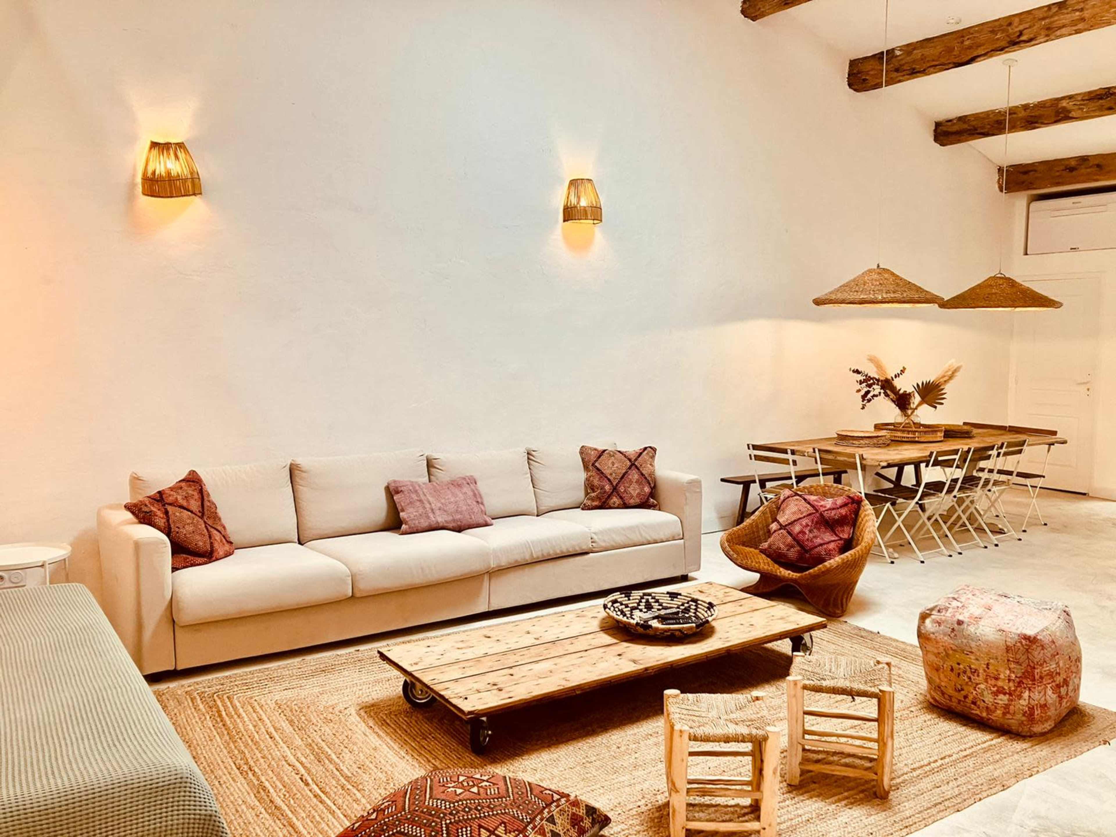The image depicts a spacious living area featuring a light-colored couch, woven light fixtures, a wooden dining table, and various decorative elements on the floor.