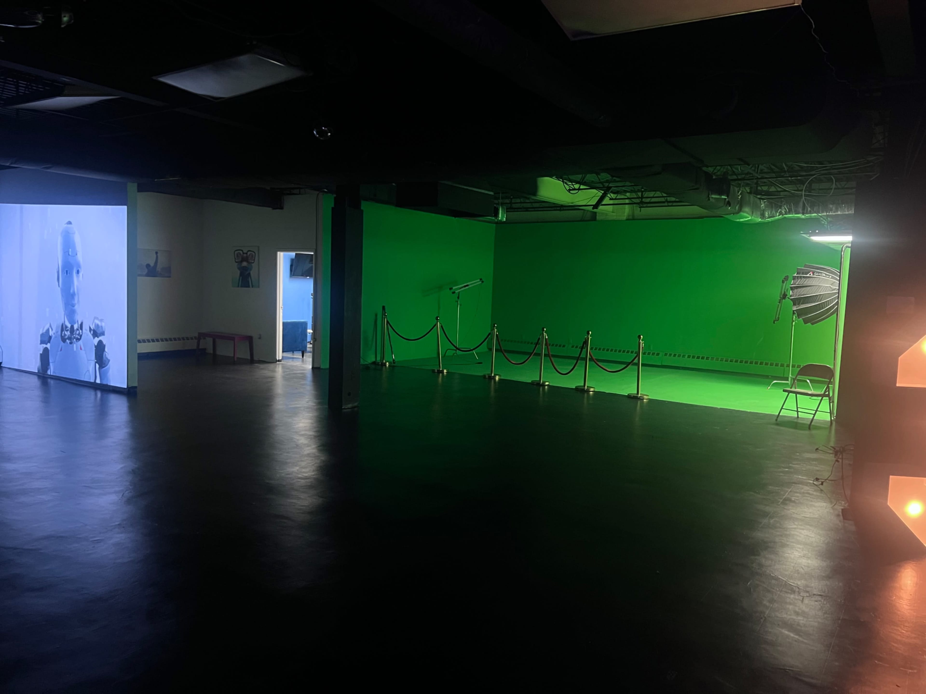 An indoor space featuring a green screen area, a wall displaying a video, and a section with ropes and a chair for audience or production use.