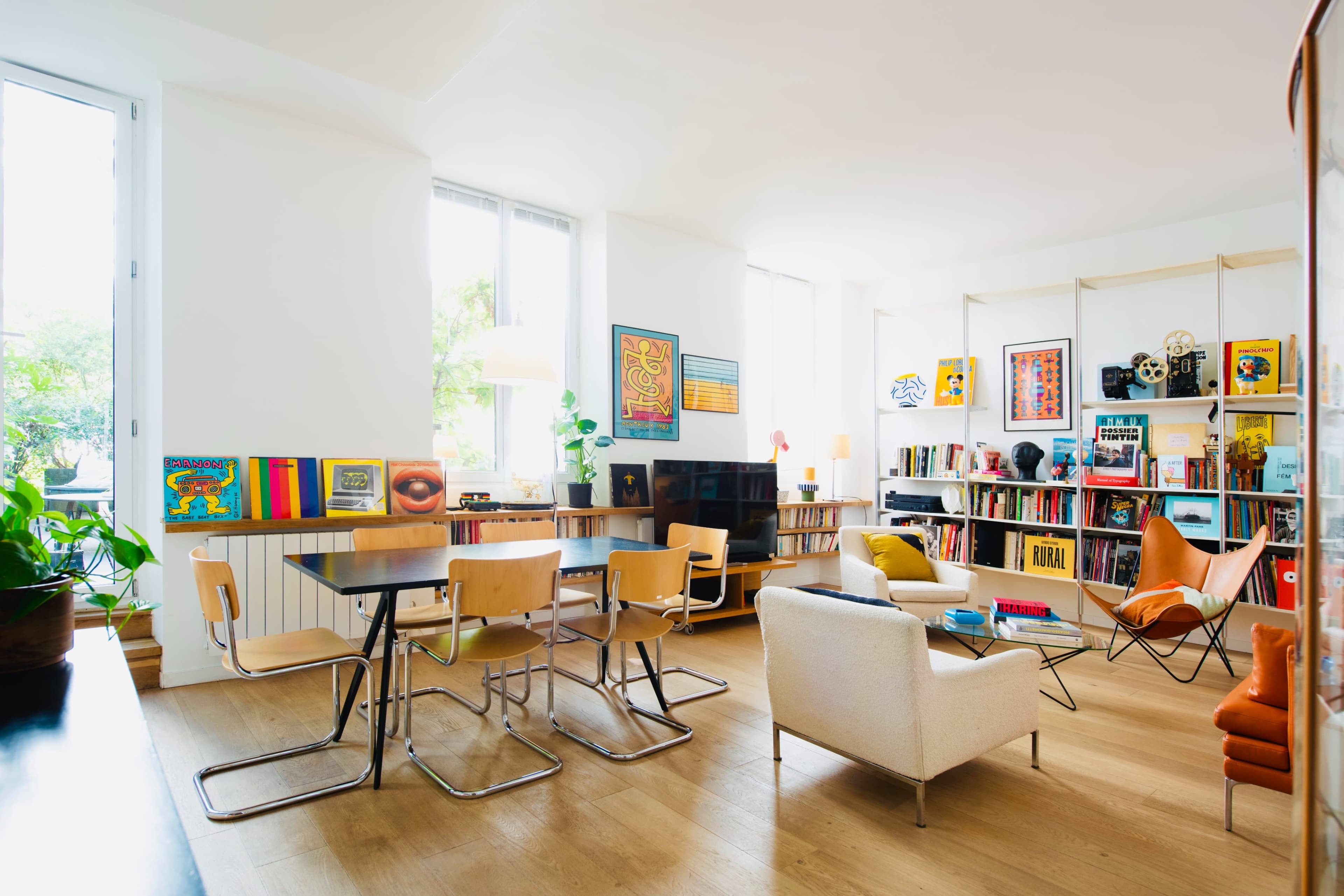 A bright living room features a dining table with four chairs, a comfortable armchair, and a bookshelf filled with various books and art pieces.