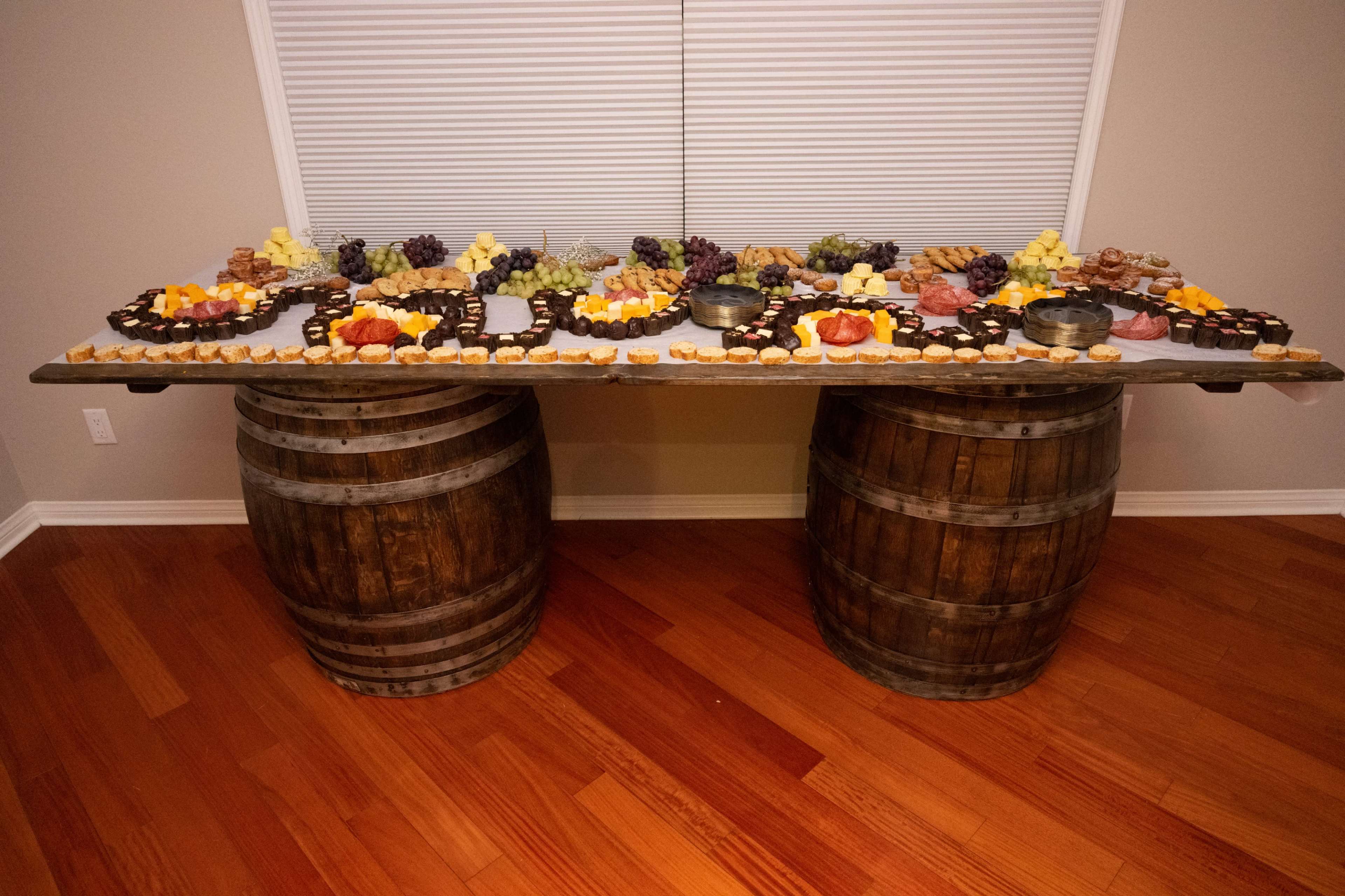 A large, multi-tiered display of assorted fruits, cheeses, and desserts is arranged on a table made from wooden barrels.