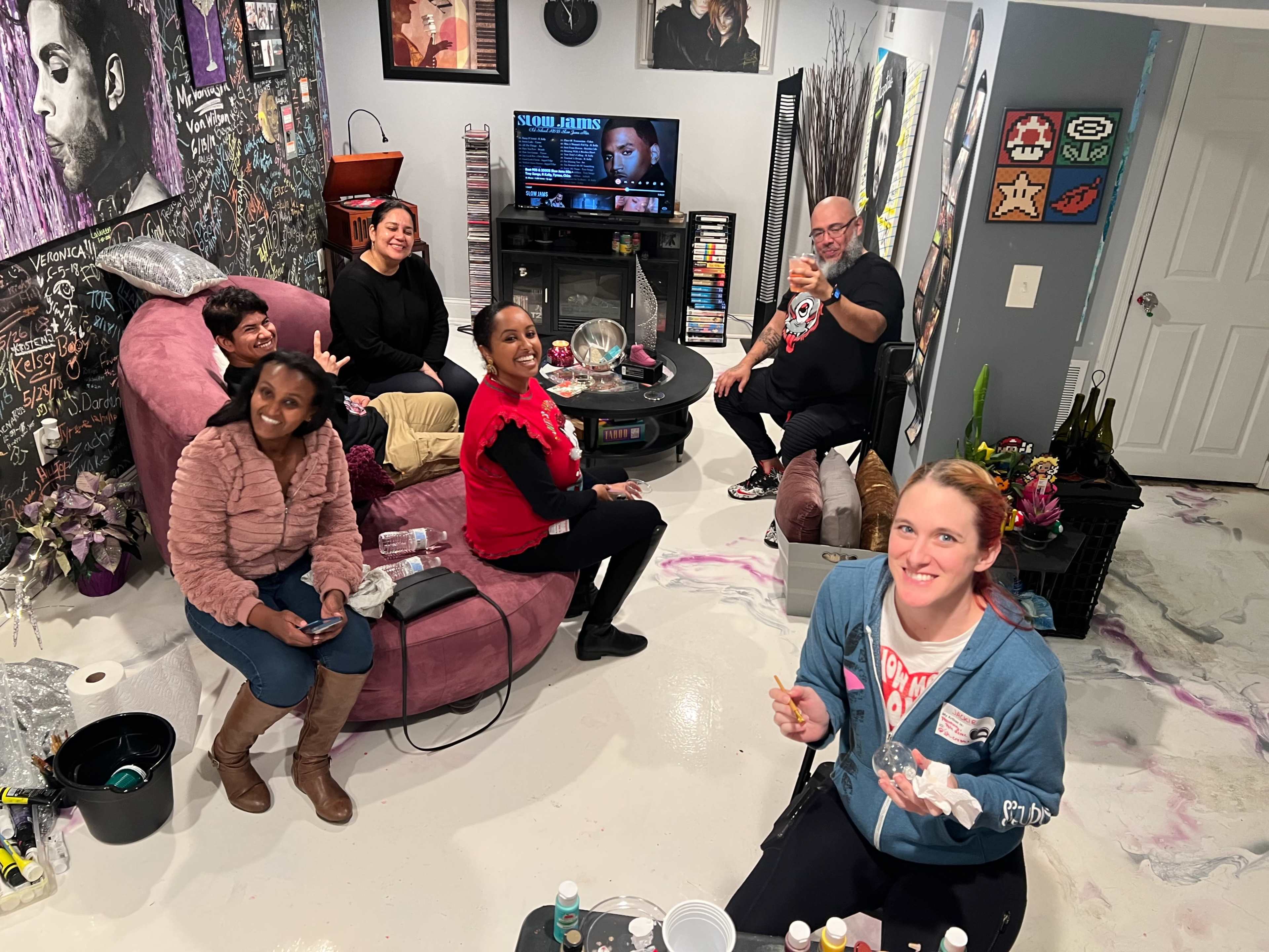 A group of six people gather in a colorful living room, engaged in a painting activity while watching a movie on TV.