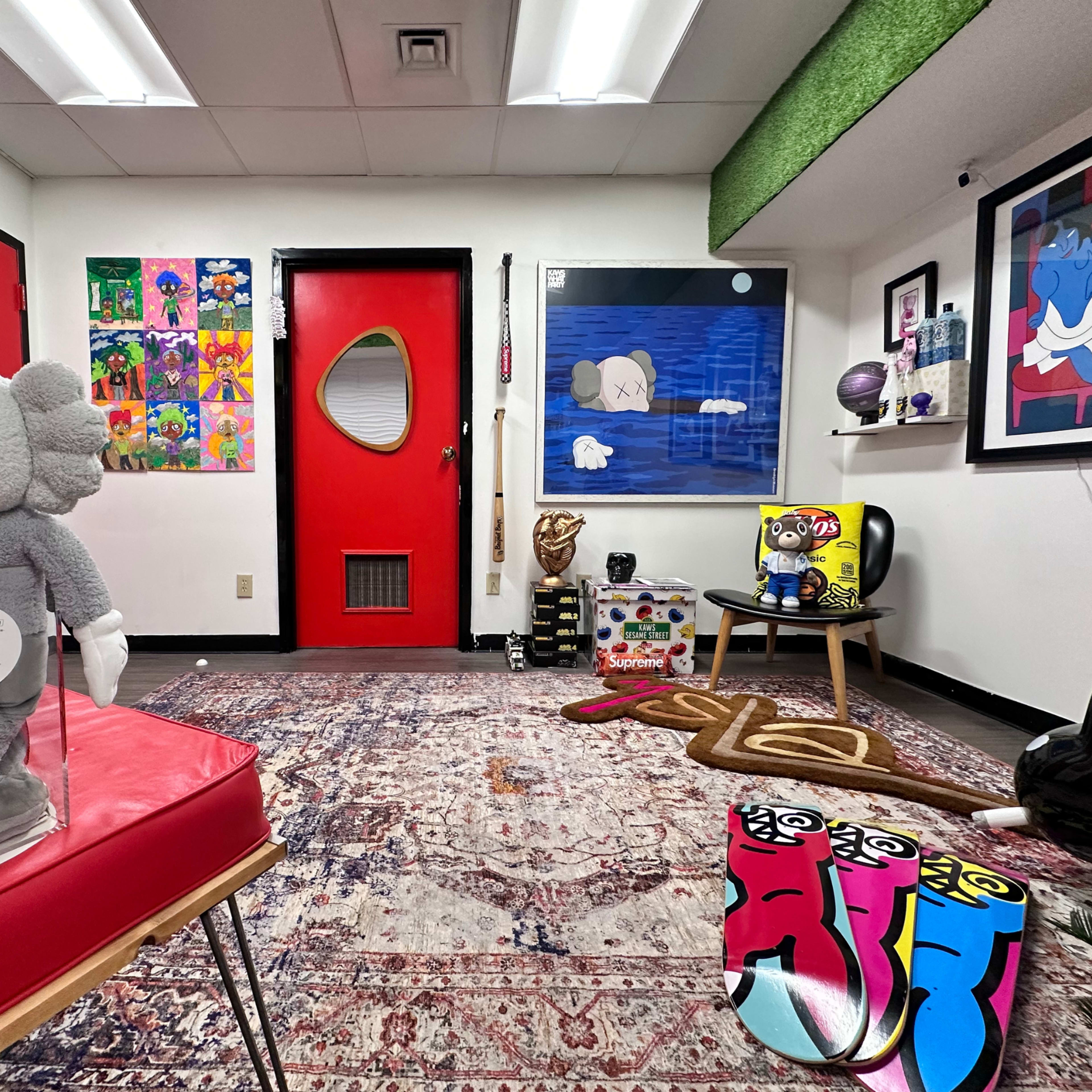 The image shows a small, art-filled room featuring colorful posters on one wall, a red door, various decorations including plush toys and art pieces, and a patterned rug on the floor.