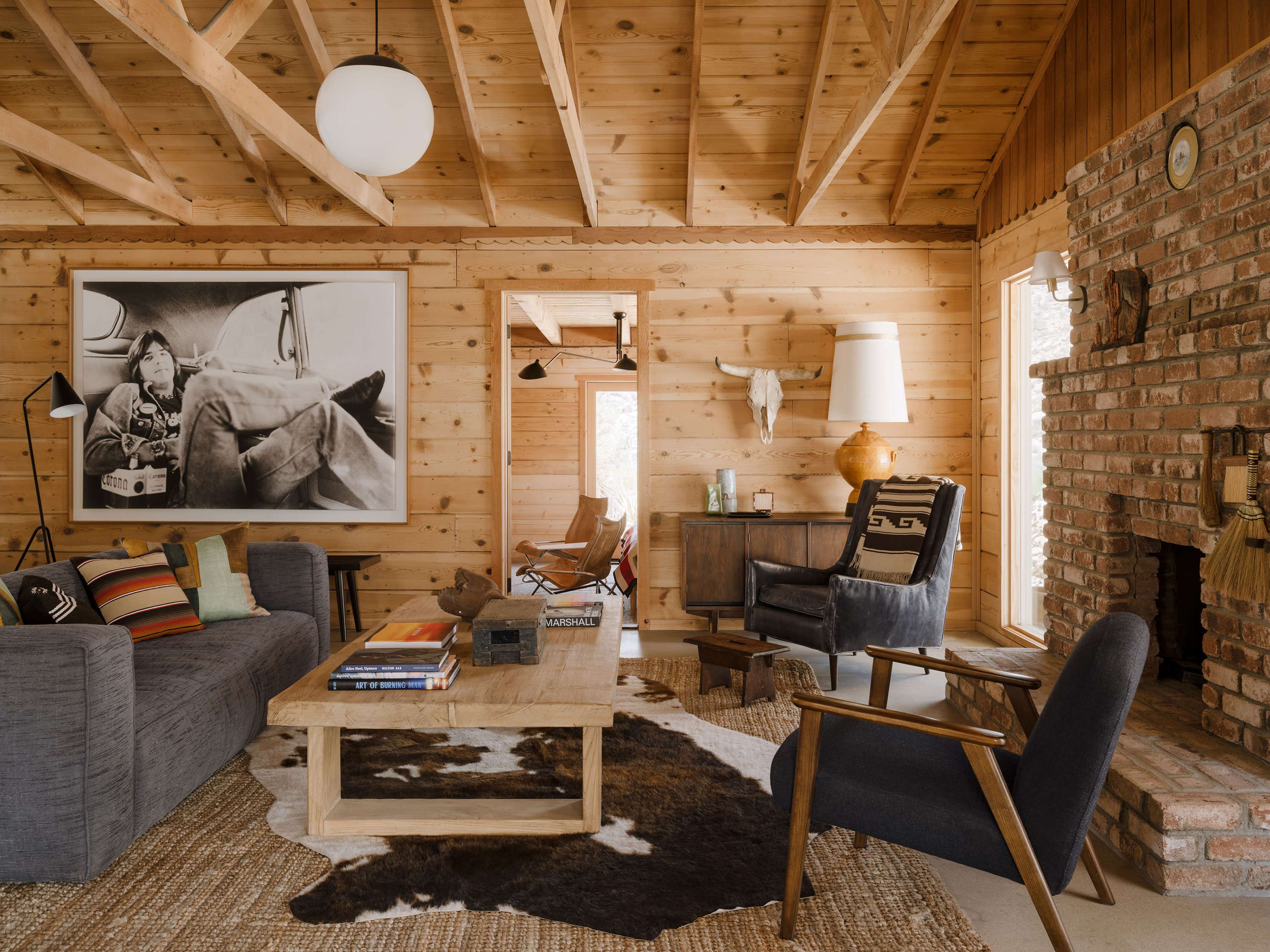 The image shows a cozy wooden cabin living room with a large photo on the wall, a coffee table on a cowhide rug, and various pieces of furniture arranged around a brick fireplace.