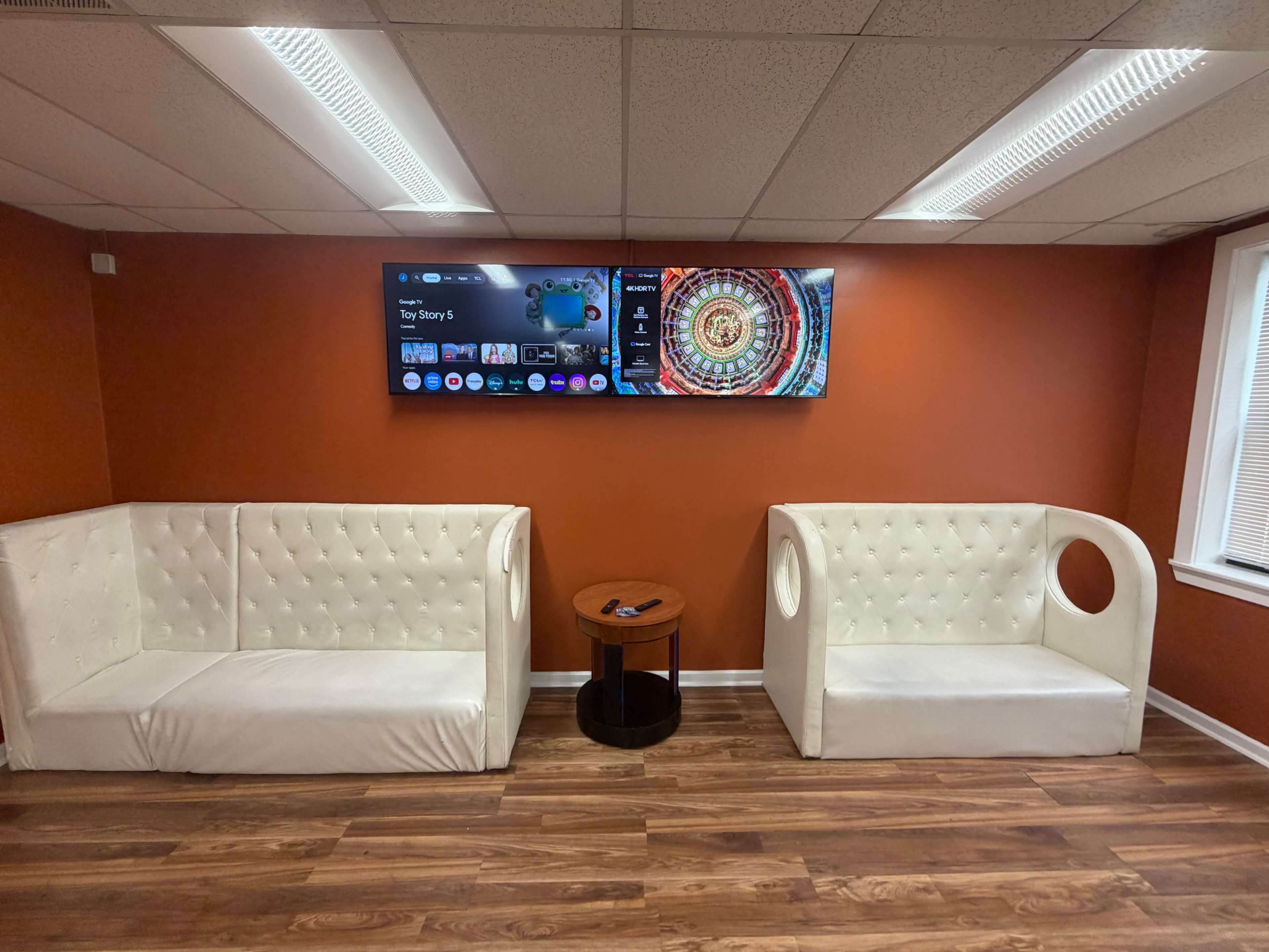 The image shows a waiting area with two white upholstered sofas and a small round wooden table, set against an orange wall featuring two flat-screen televisions mounted above.
