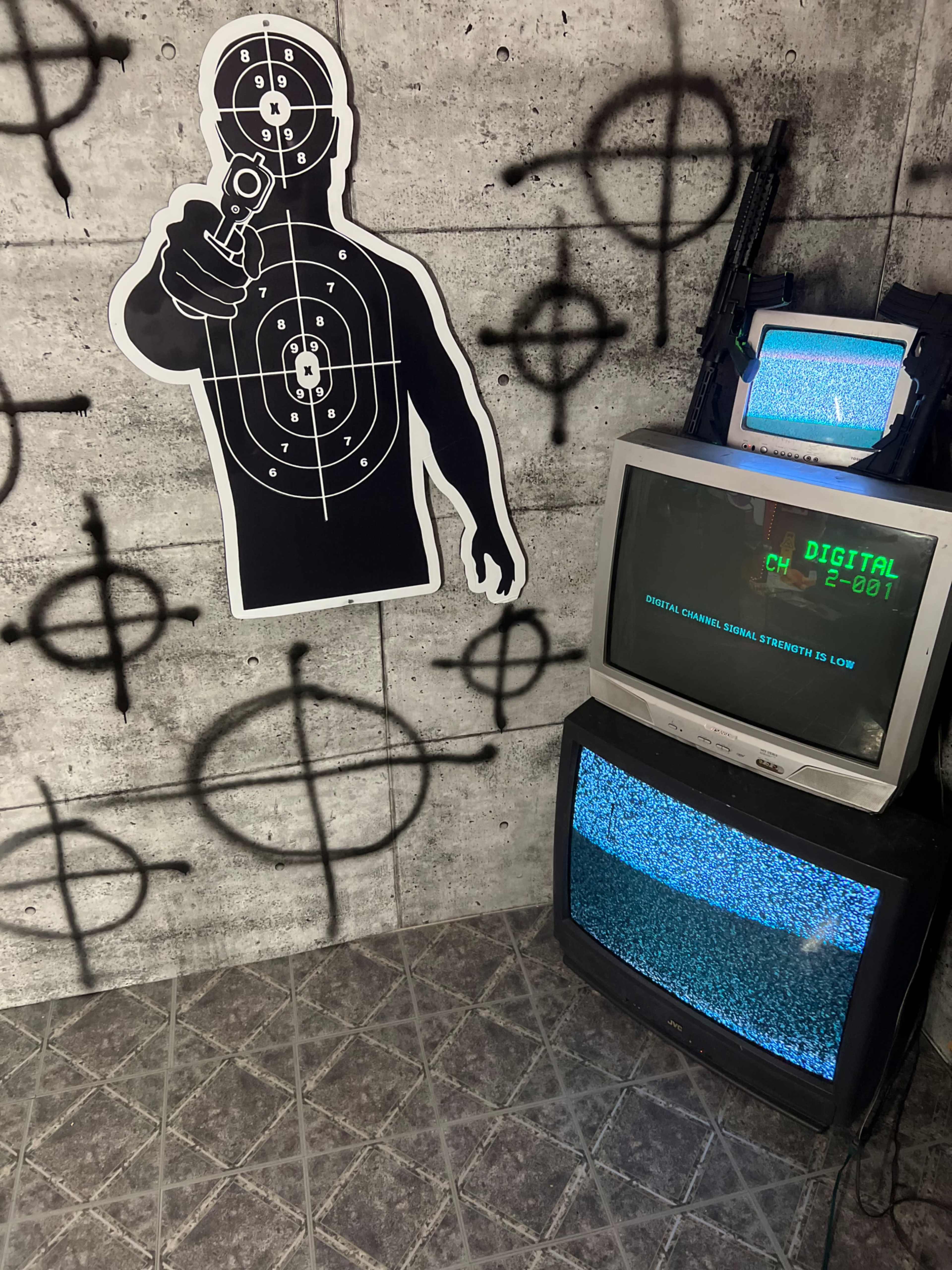 The image shows a corner with a graphic of a shooter on the wall, two old television sets displaying static, and various symbols sprayed as graffiti on a concrete background.
