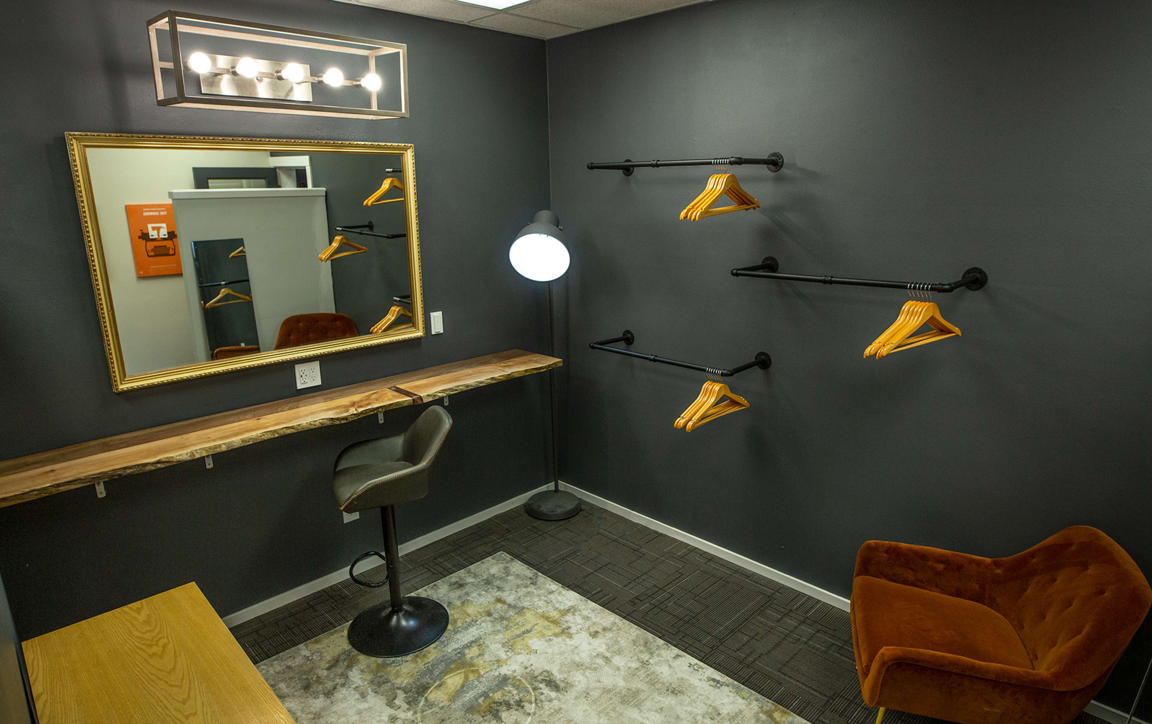 The image shows a small, modern dressing room with a large mirror, a wooden countertop, a black lamp, clothing hangers on the wall, and an orange chair.