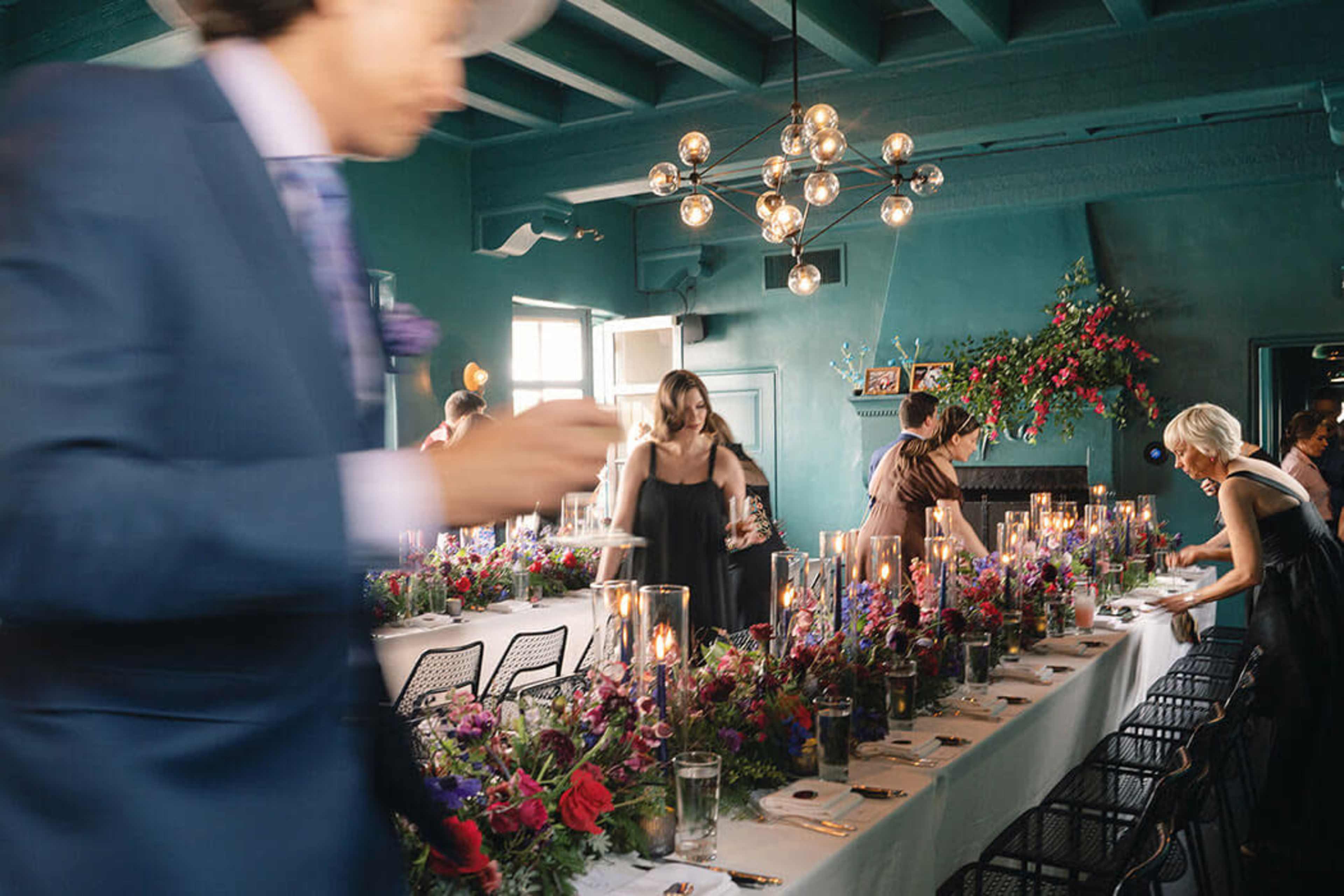 A festive dining setup features a long table adorned with flowers and candles, while guests are mingling in a warmly decorated room.