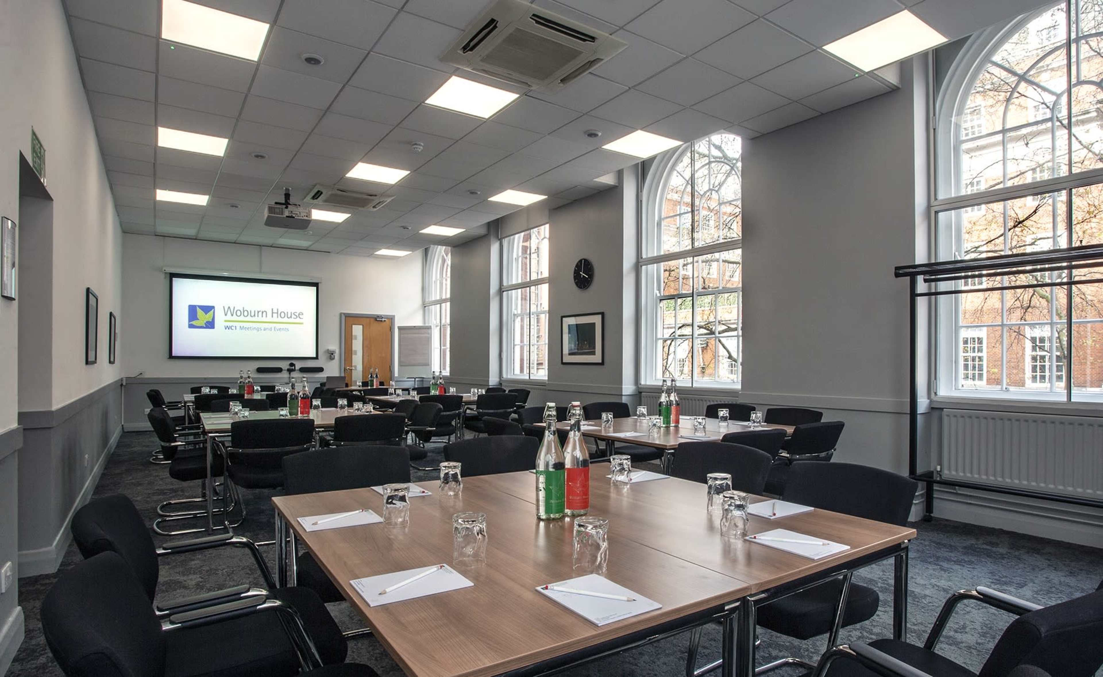 The image shows a conference room set up with several tables arranged for a meeting, featuring chairs and water bottles on each table, with a screen displaying "Woburn House" in the background.
