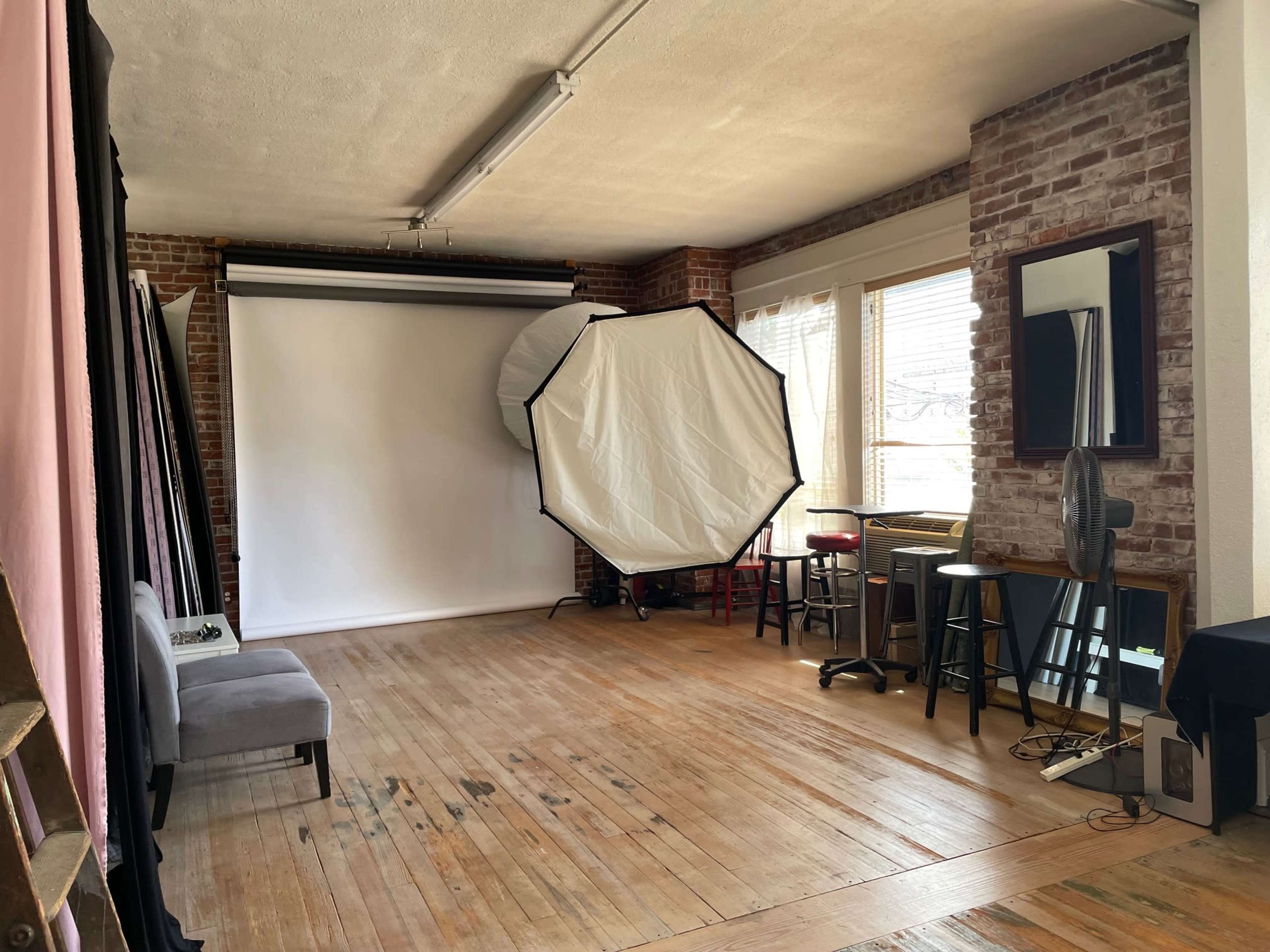 A spacious photography studio with brick walls, wooden flooring, and a backdrop stand featuring a large white screen.