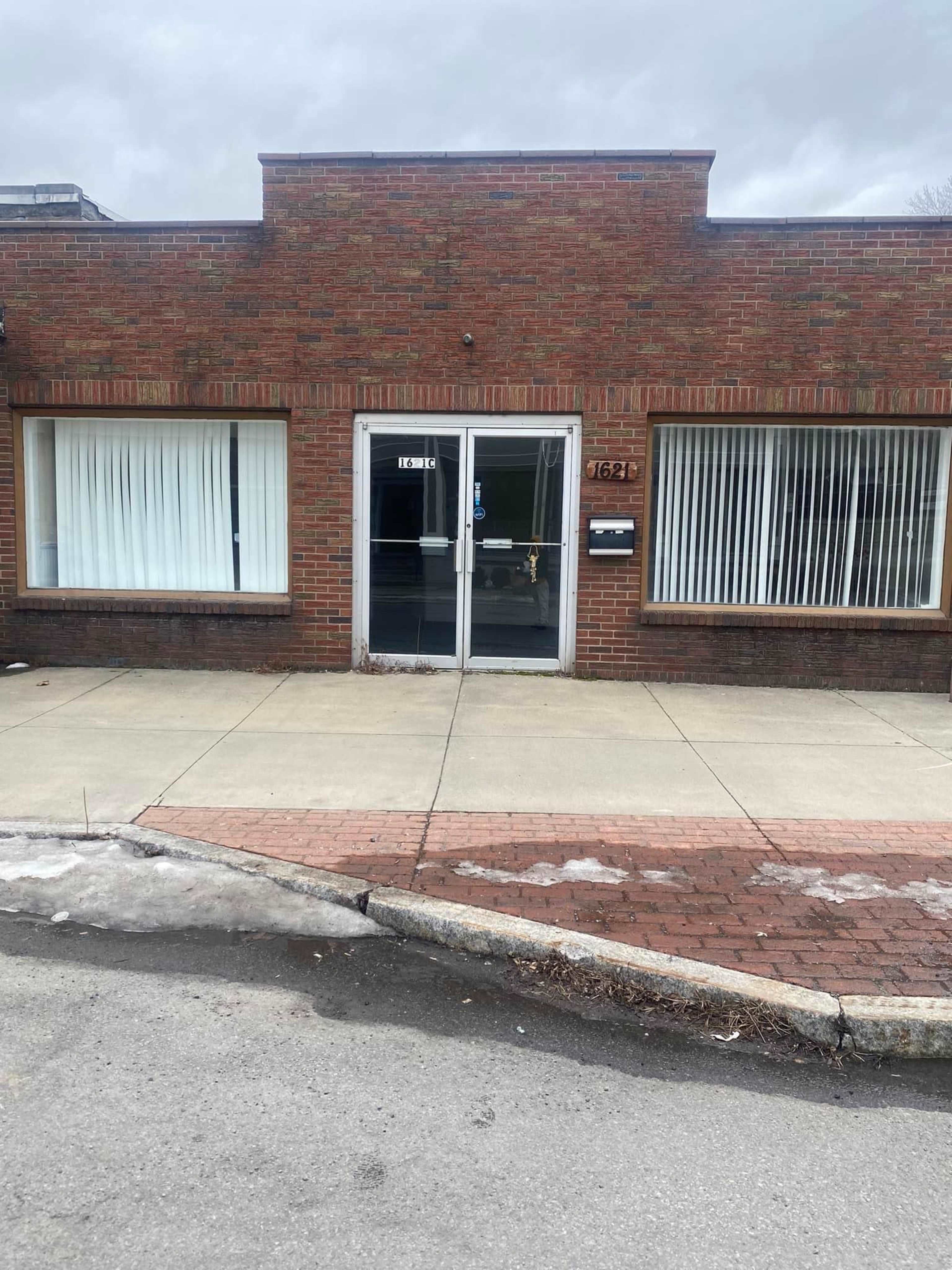 A brick building with two large windows and a central glass door, located on a pavement beside a road.