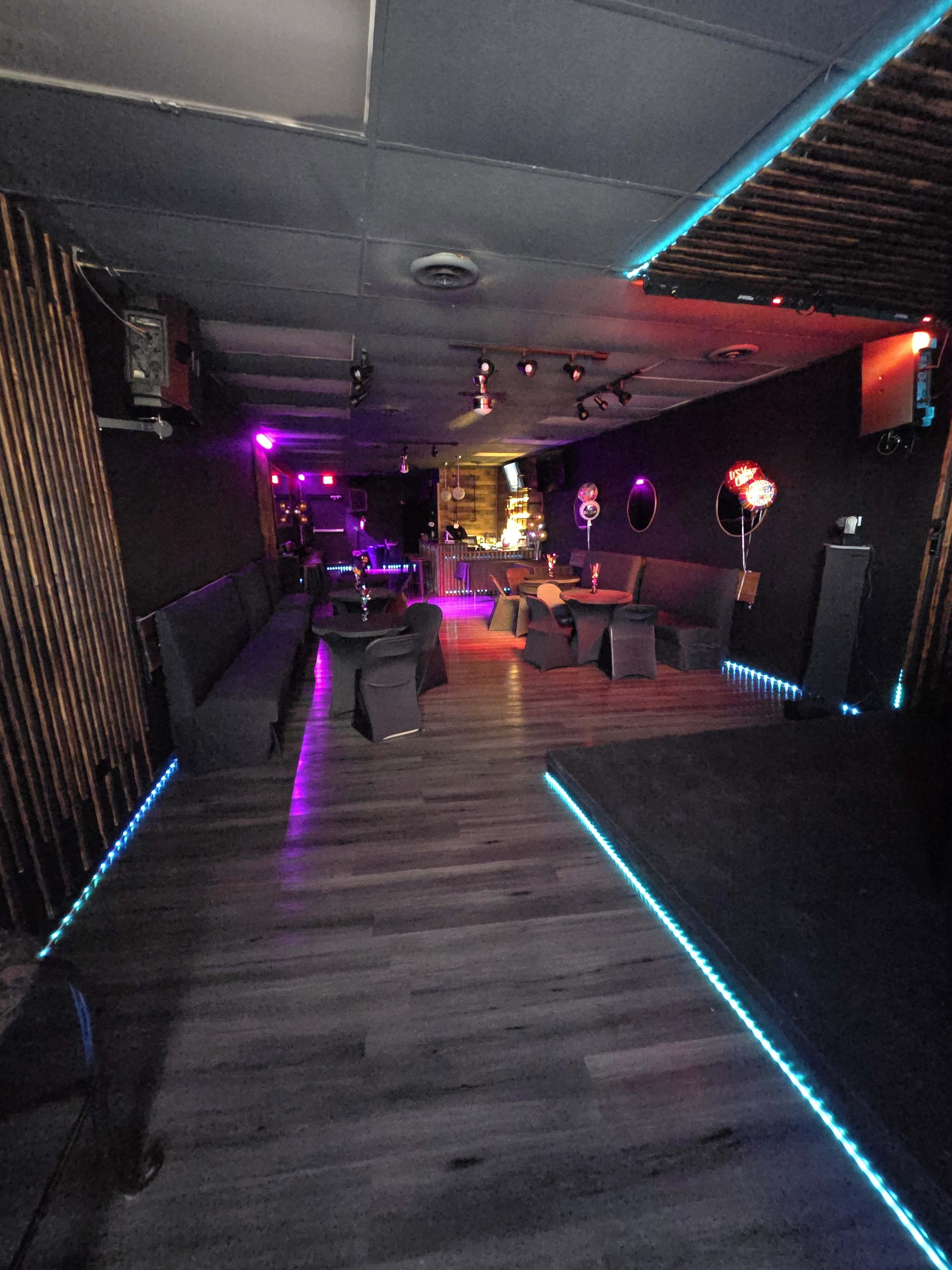 The image shows the interior of a dimly lit lounge with seating arrangements and colorful ambient lighting along the floors.