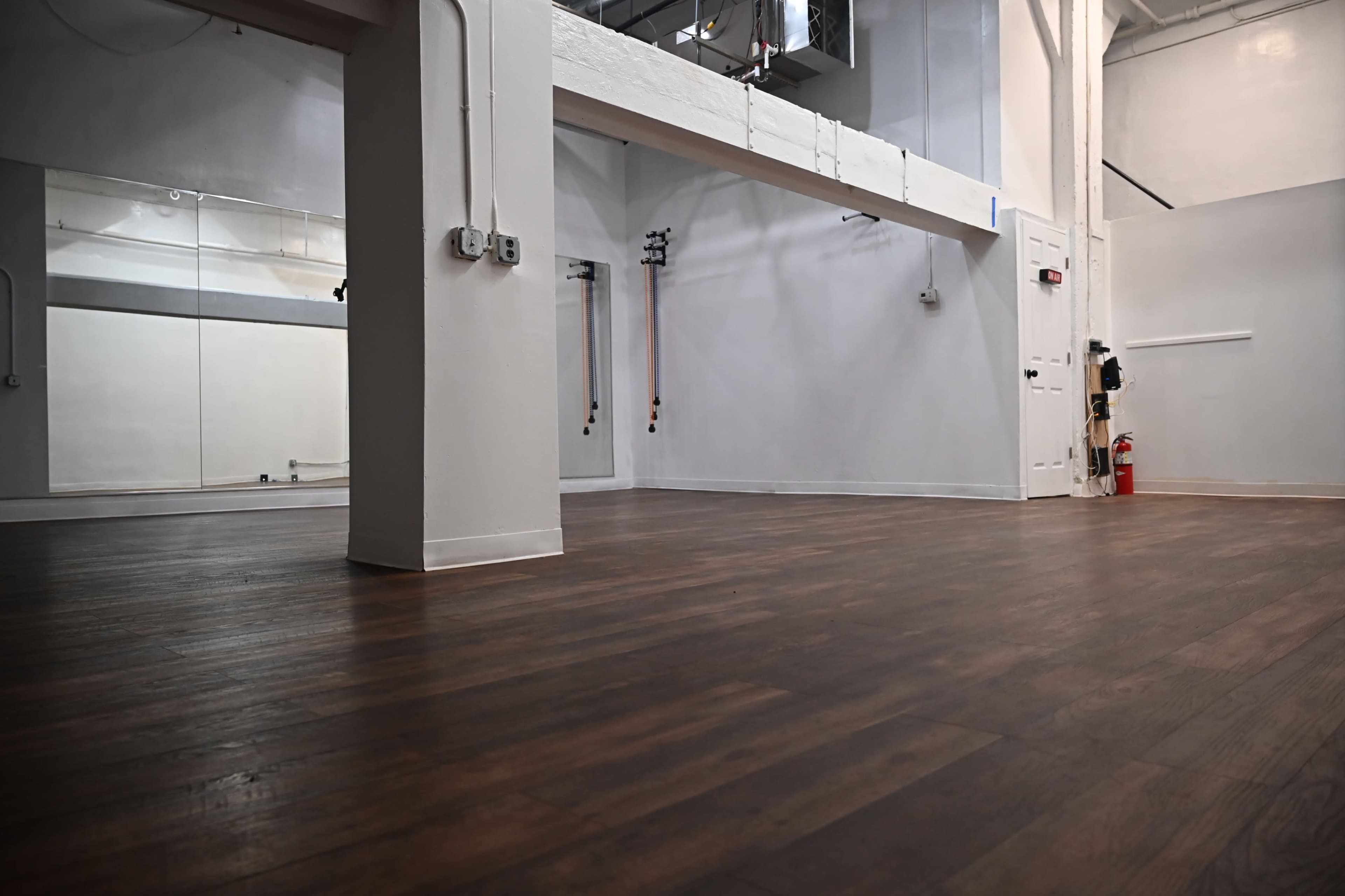 An empty, well-lit room with wooden flooring and white walls, featuring large mirrors and a few wall-mounted equipment.