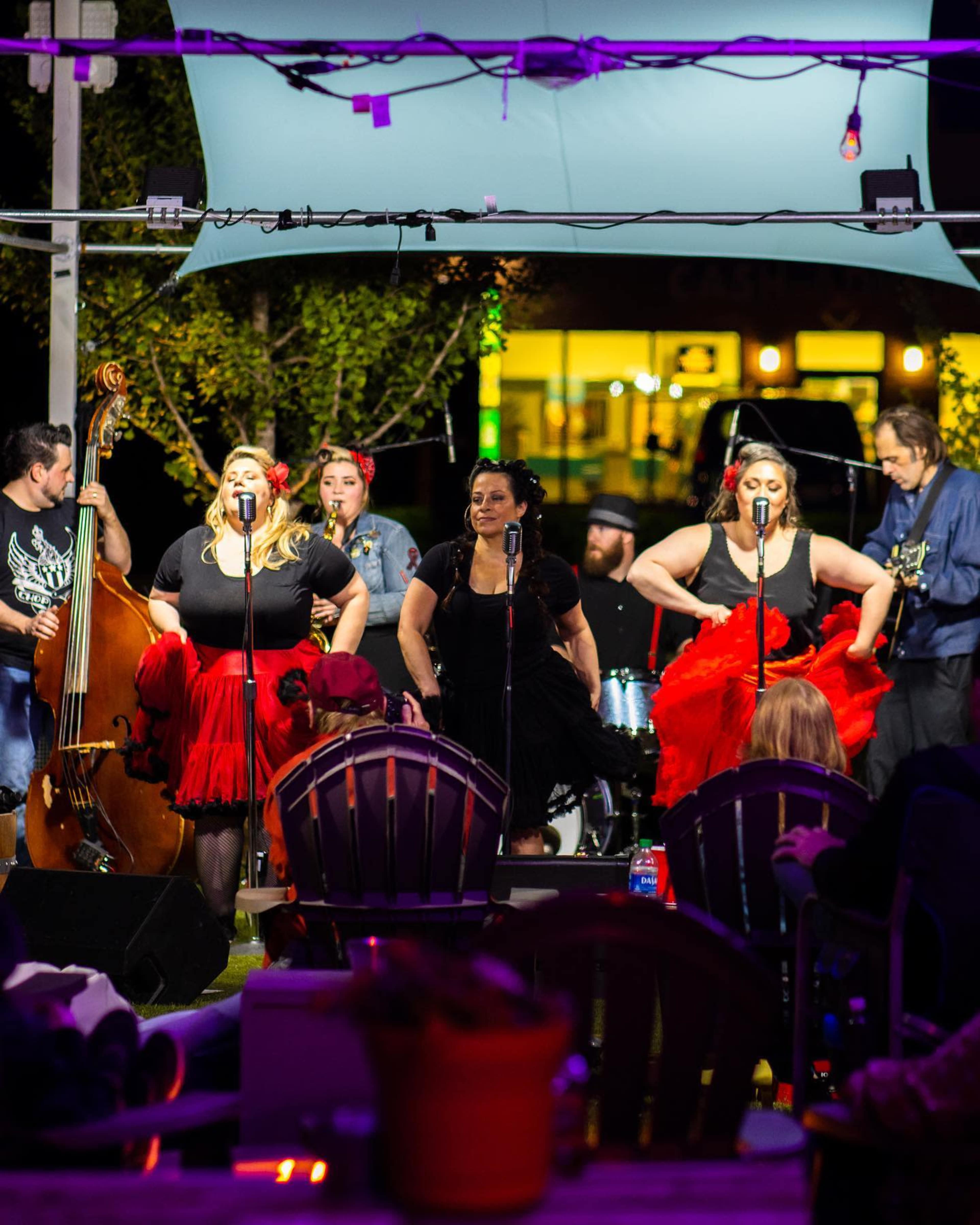 A group of performers in black and red outfits is singing into microphones while a band plays behind them on an outdoor stage.