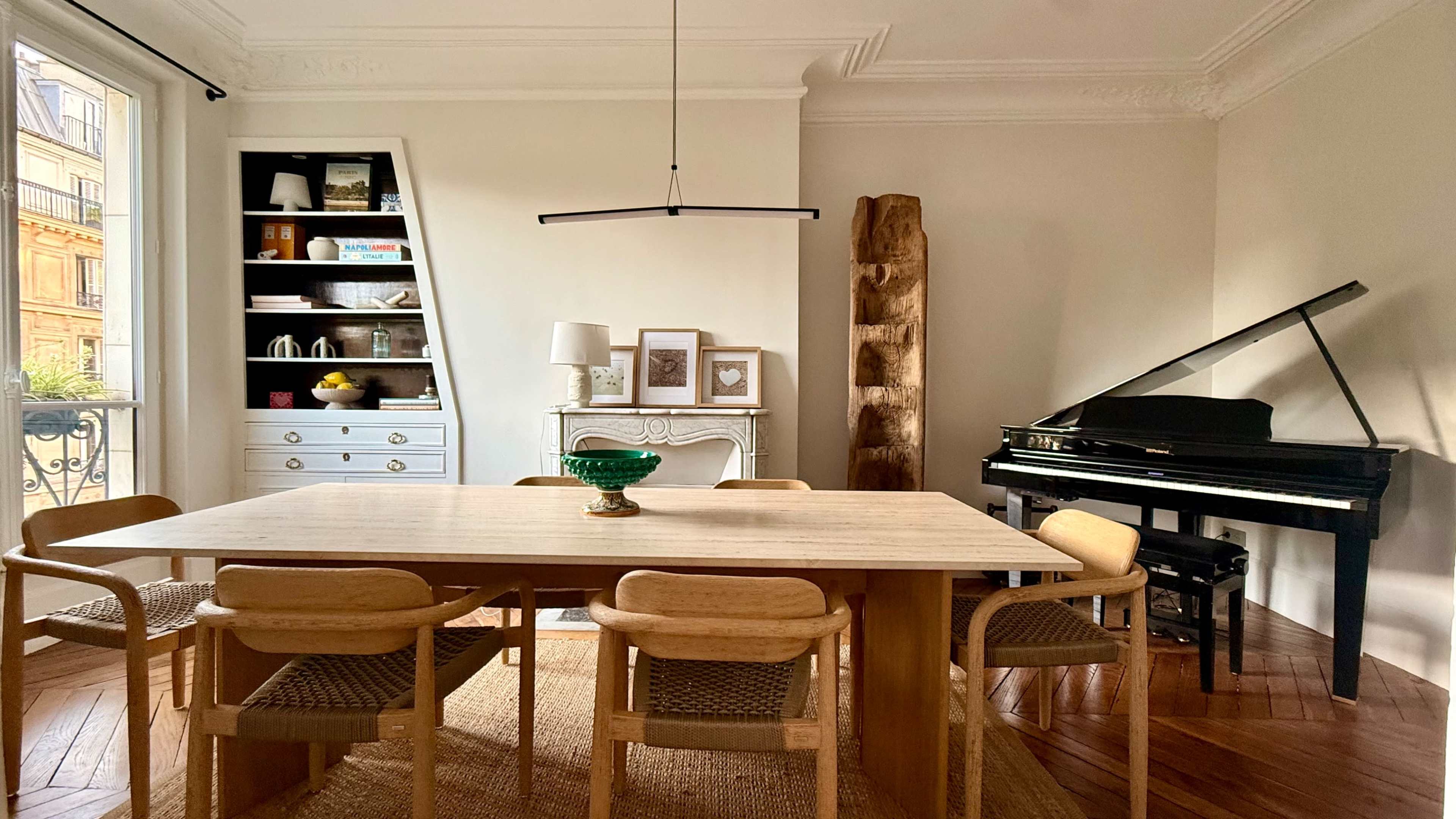 The image depicts a well-lit dining area featuring a wooden table surrounded by six chairs, a black piano in the corner, and shelves displaying various decorative items.