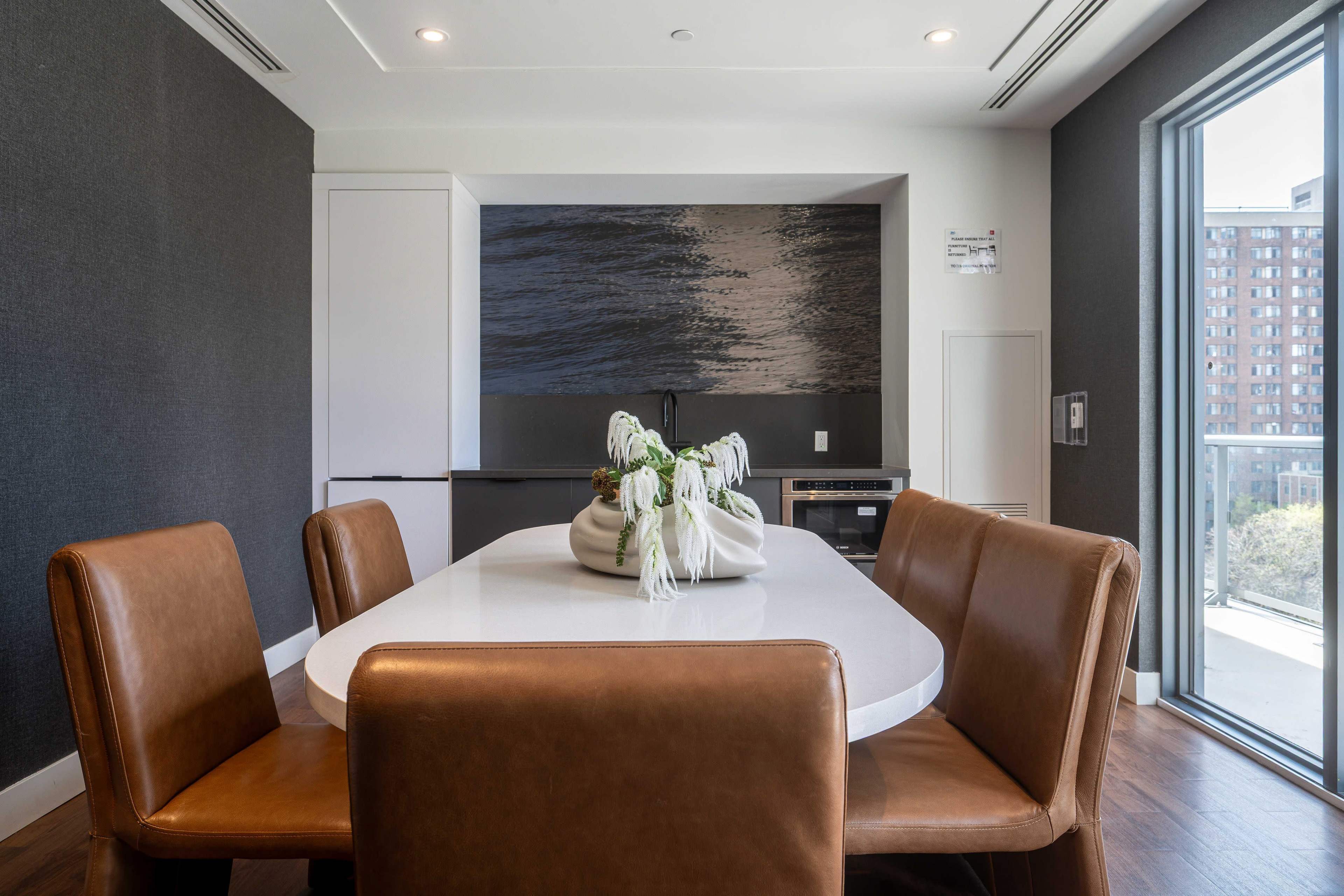 A modern dining area features a white table surrounded by brown leather chairs, with a decorative vase at the center and a view of a cityscape through the glass door.