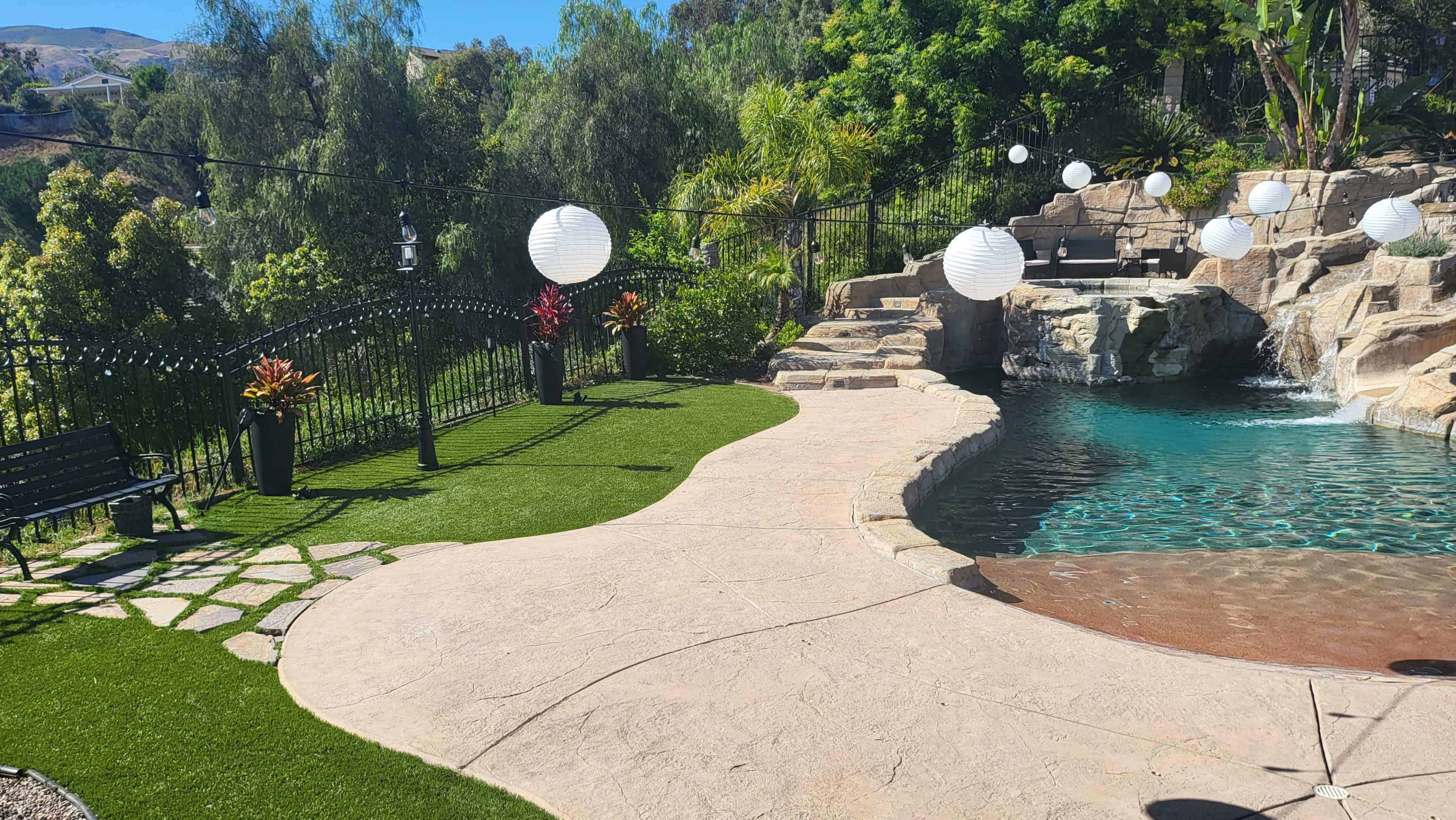 A landscaped backyard featuring a pool, stone steps, green grass, and decorative lanterns hanging above.