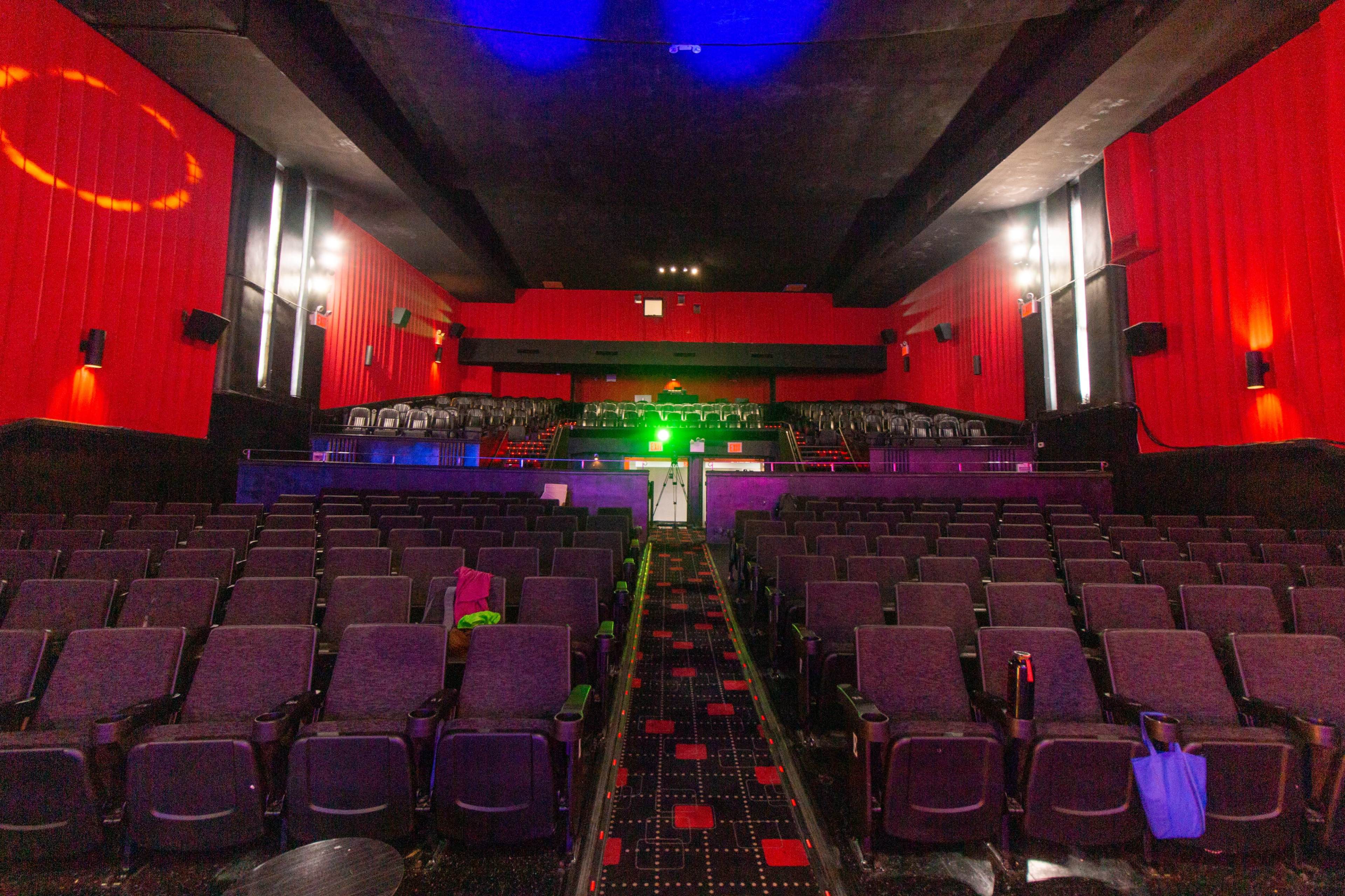 An empty movie theater with red walls, several rows of black seats, and a stage illuminated by green light.