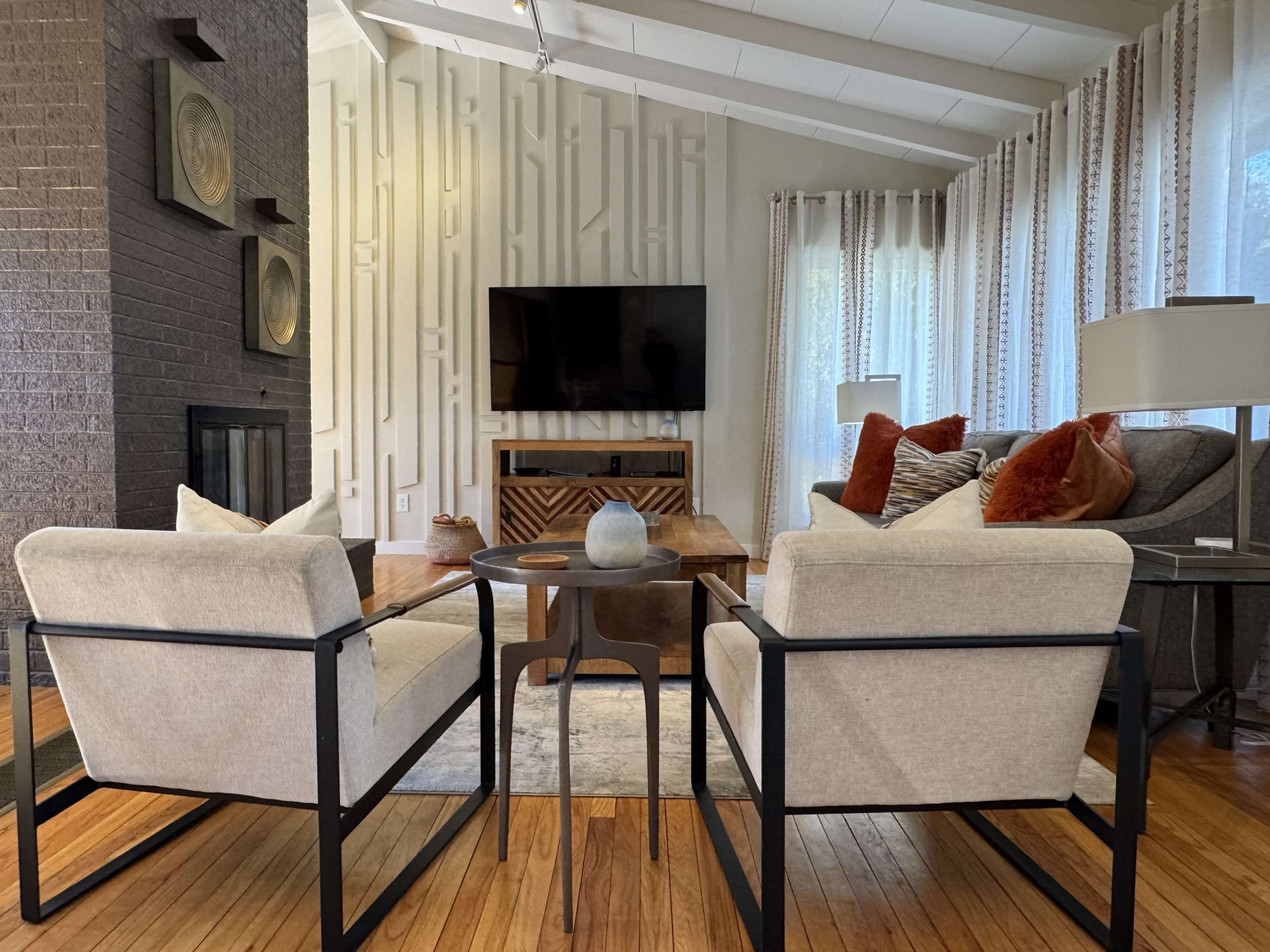 The image shows a cozy living room featuring two armchairs, a wooden coffee table, a TV mounted on the wall, and decorative curtains.