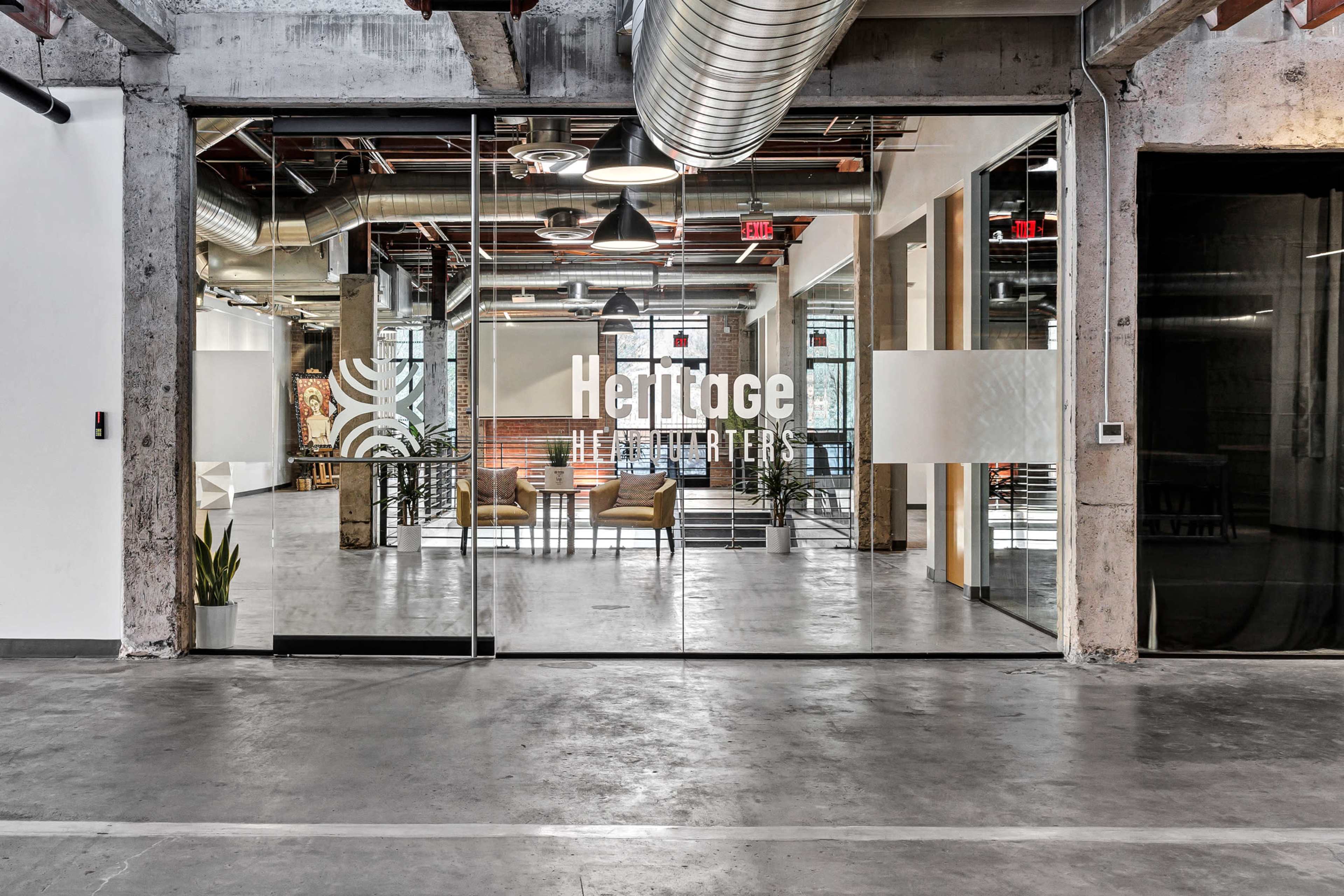 The image shows a modern office space with a glass entrance featuring the text "Heritage Headquarters" prominently displayed.