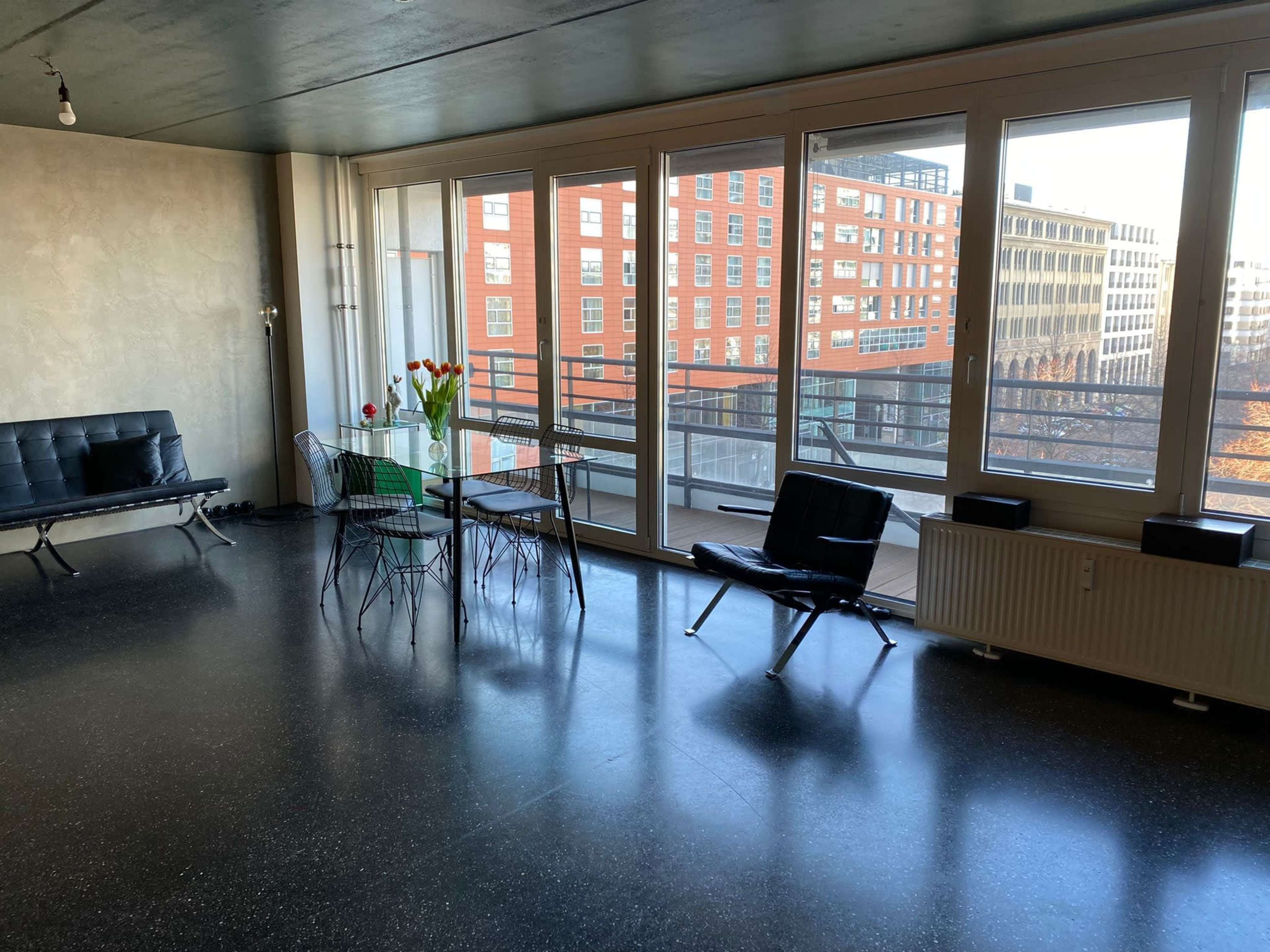 The image shows a modern interior space with large windows, a dining table with chairs, and a black sofa.