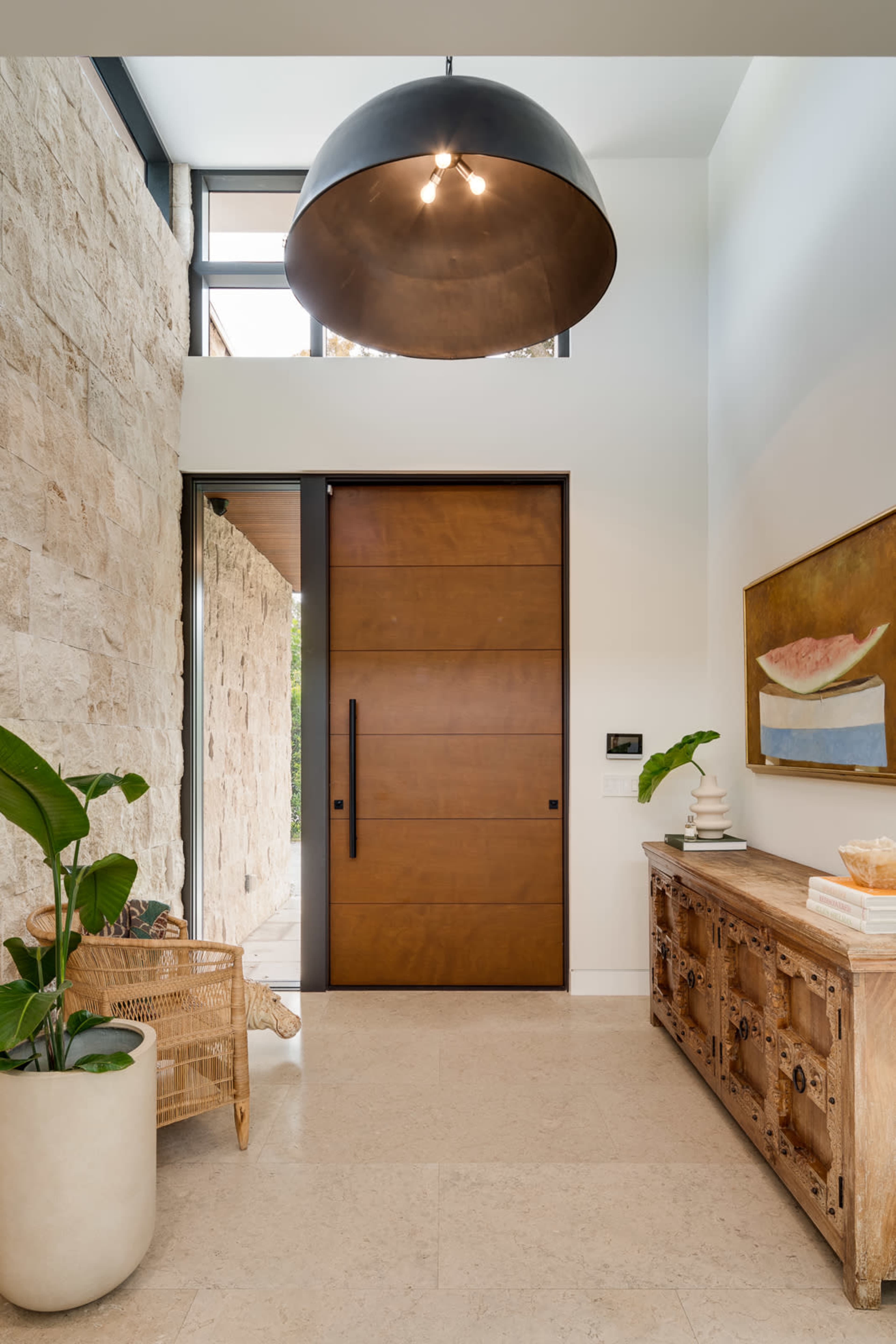 A modern entryway featuring a large wooden door, natural stone wall, and a decorative wooden console table beside a potted plant.
