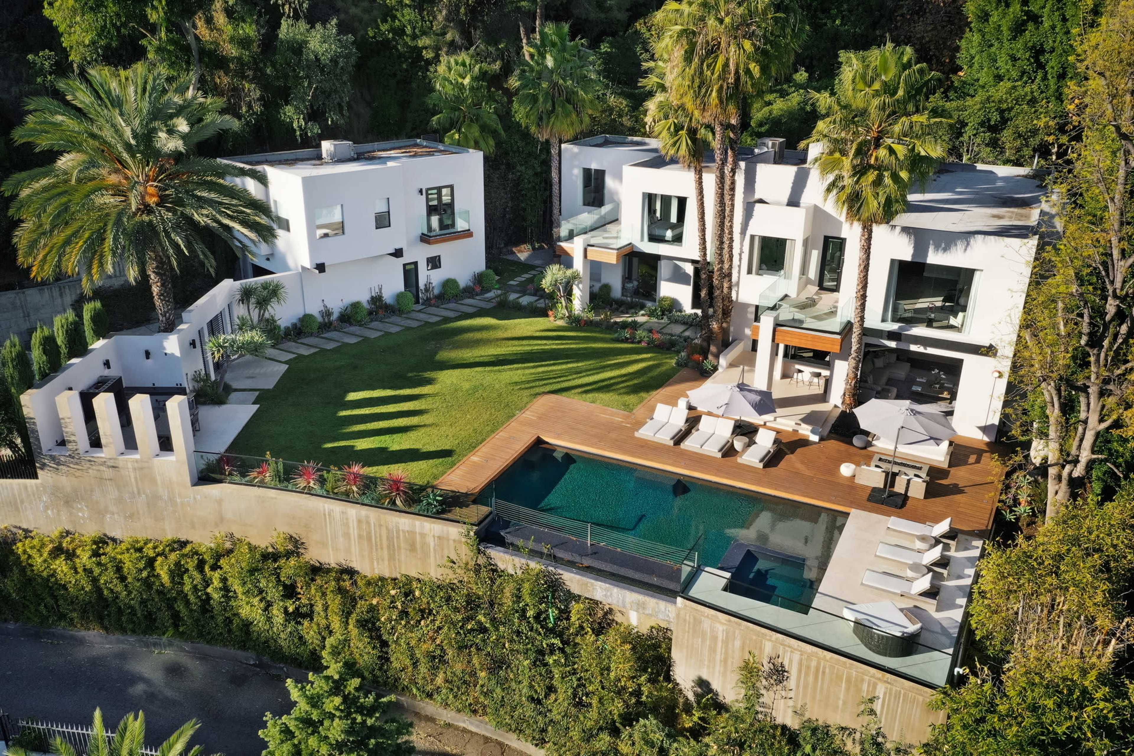 The image shows a modern two-story house with a grassy lawn, palm trees, and a swimming pool surrounded by a wooden deck.
