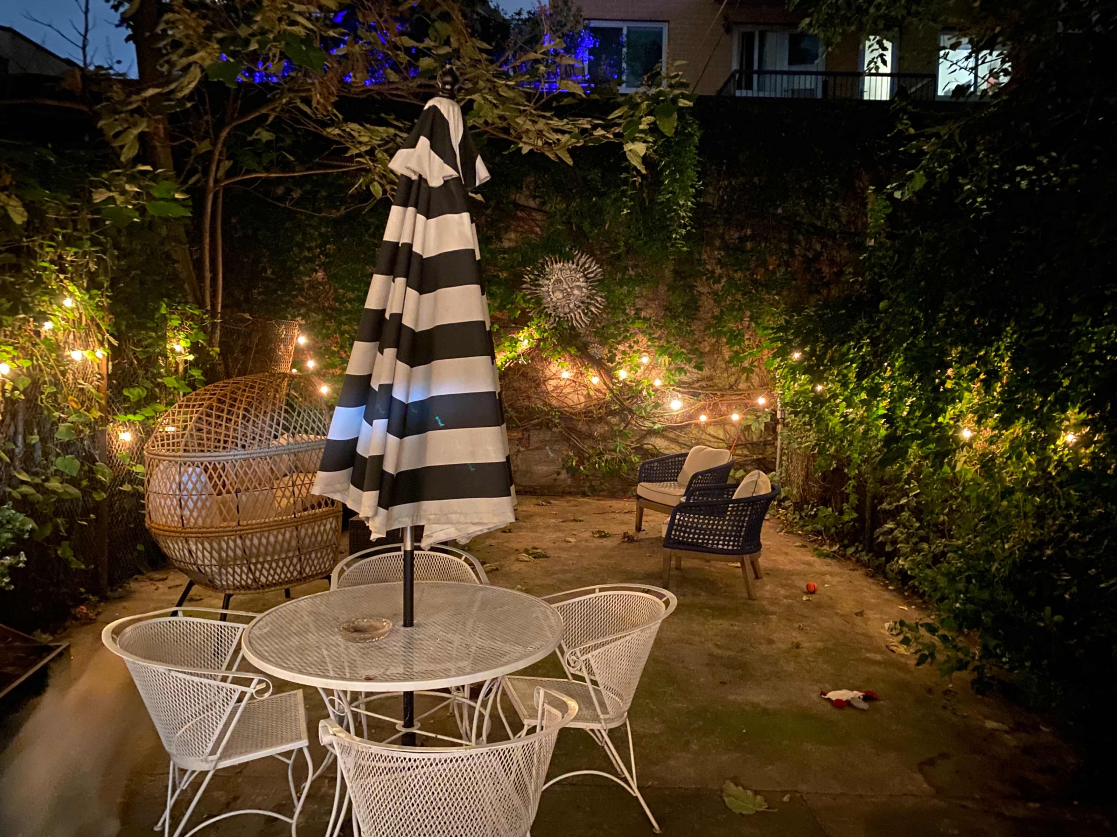 A small outdoor patio features a round table with white chairs, a striped umbrella, and string lights illuminating the space.