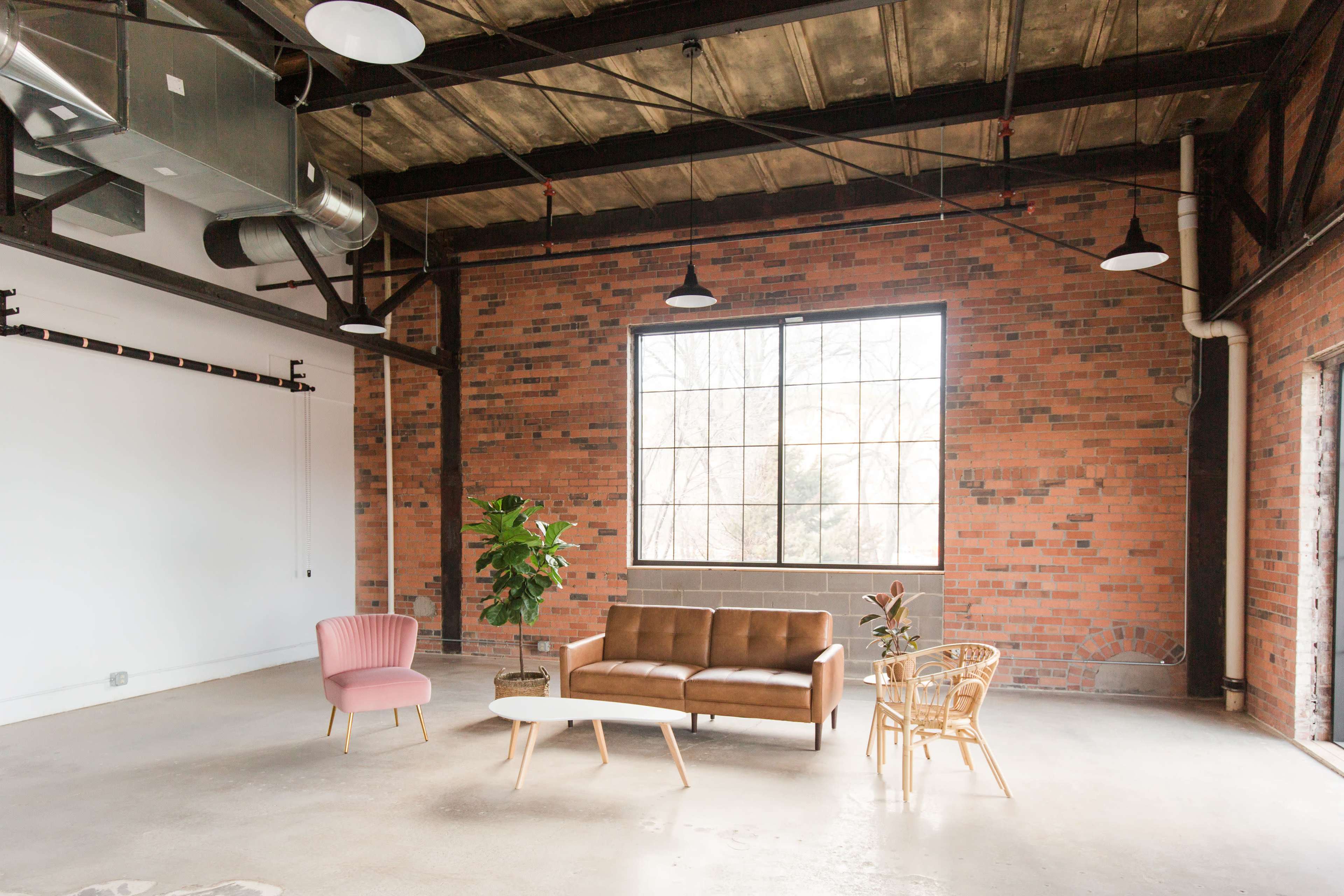 A spacious room with a large window, featuring a brown sofa, a pink armchair, a small coffee table, and a potted plant, all against a backdrop of exposed brick walls and industrial-style elements.