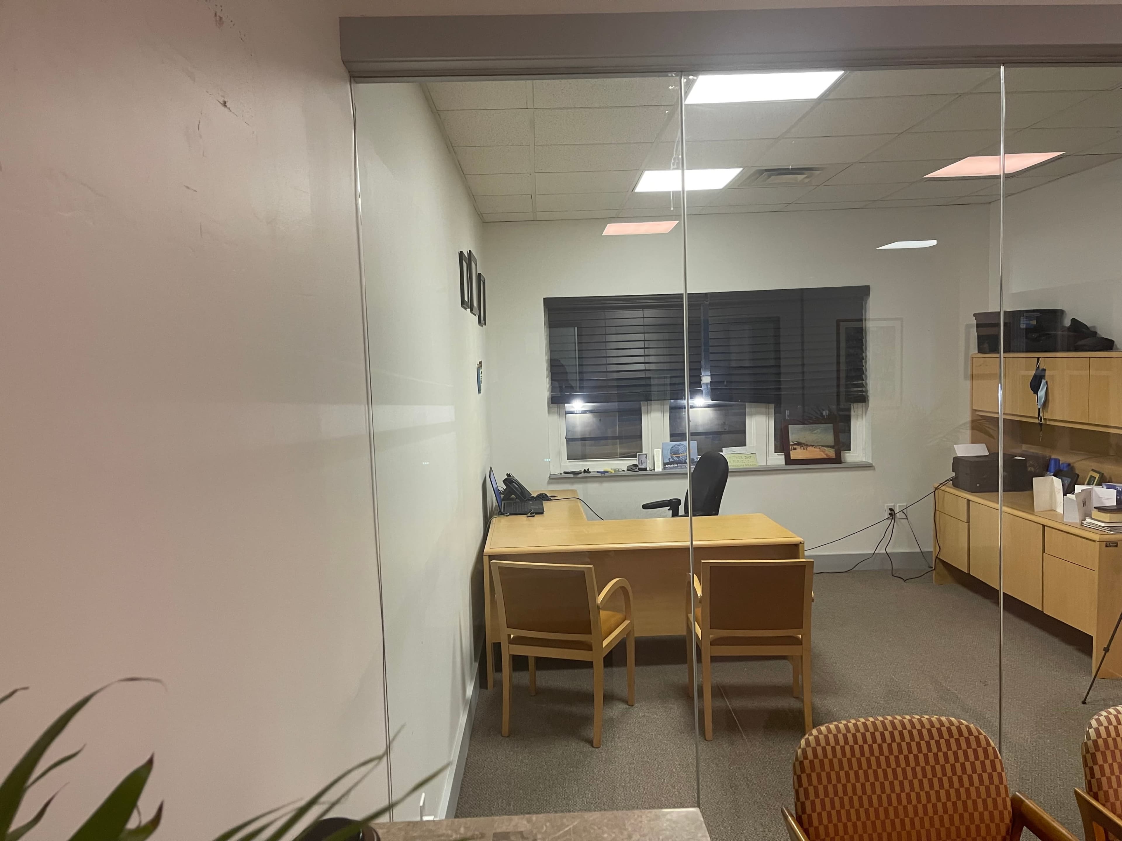A modern office space with a wooden desk and an office chair, situated behind glass walls, featuring minimal decor and large windows.