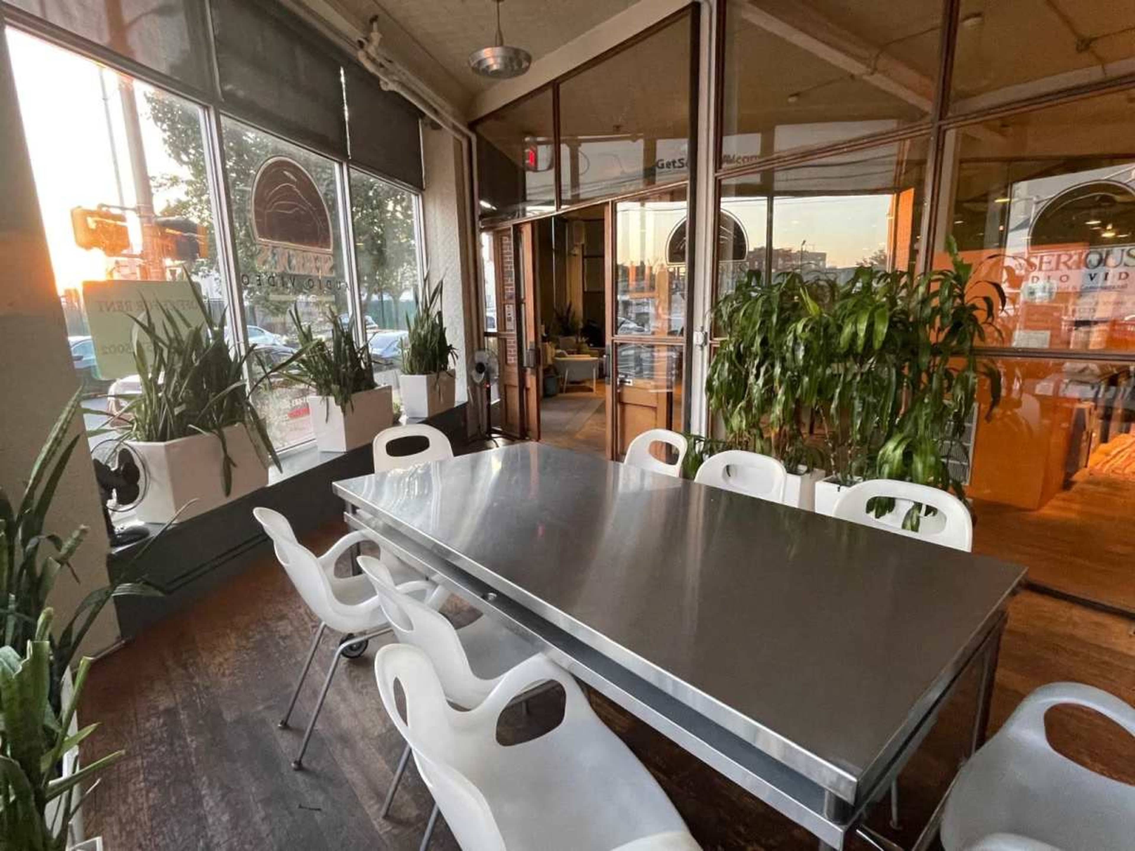 A large metal dining table is surrounded by white chairs in a sunlit room with large windows and potted plants.