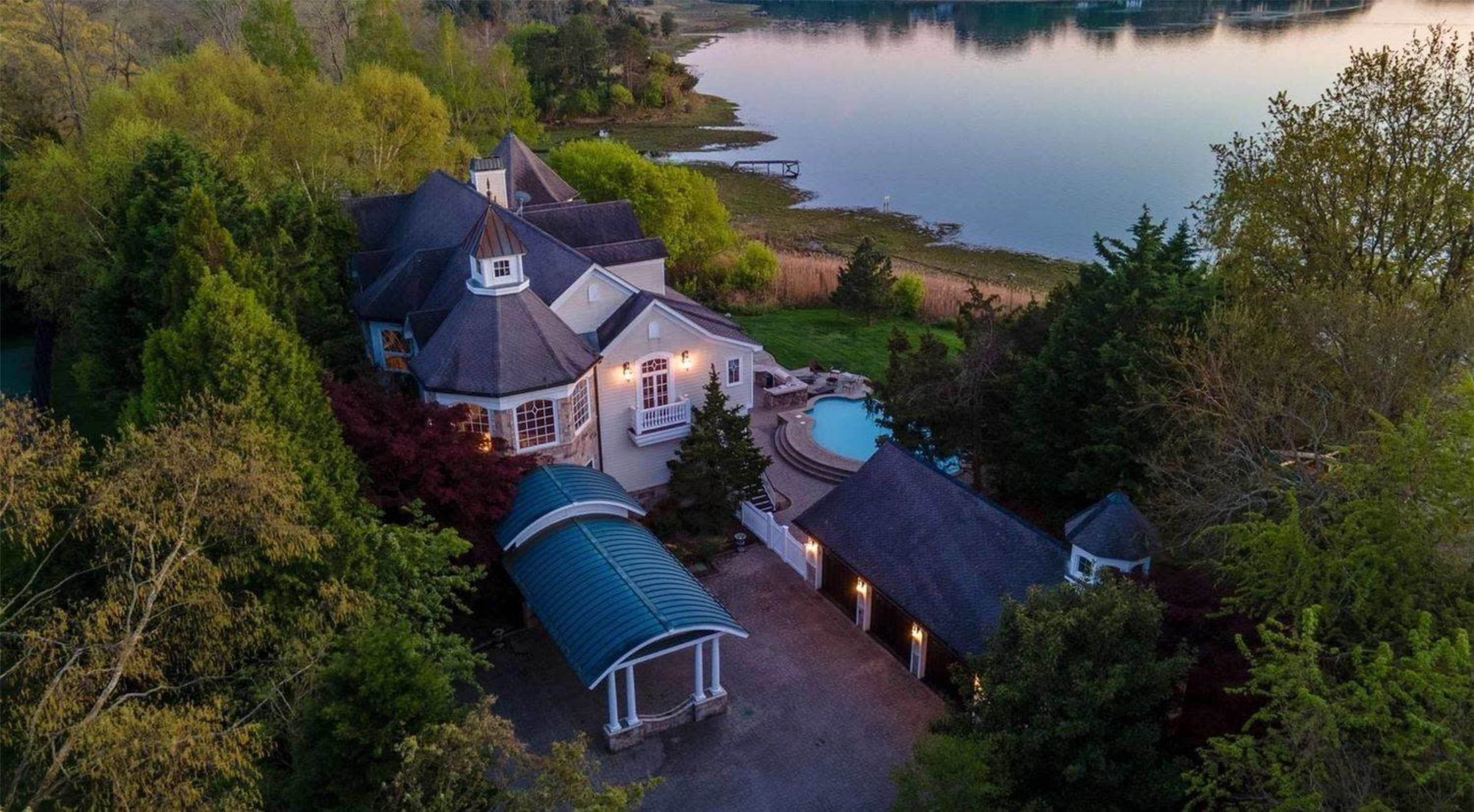 The image shows a large, multi-story house with a circular turret, a swimming pool in the backyard, and a serene body of water nearby.