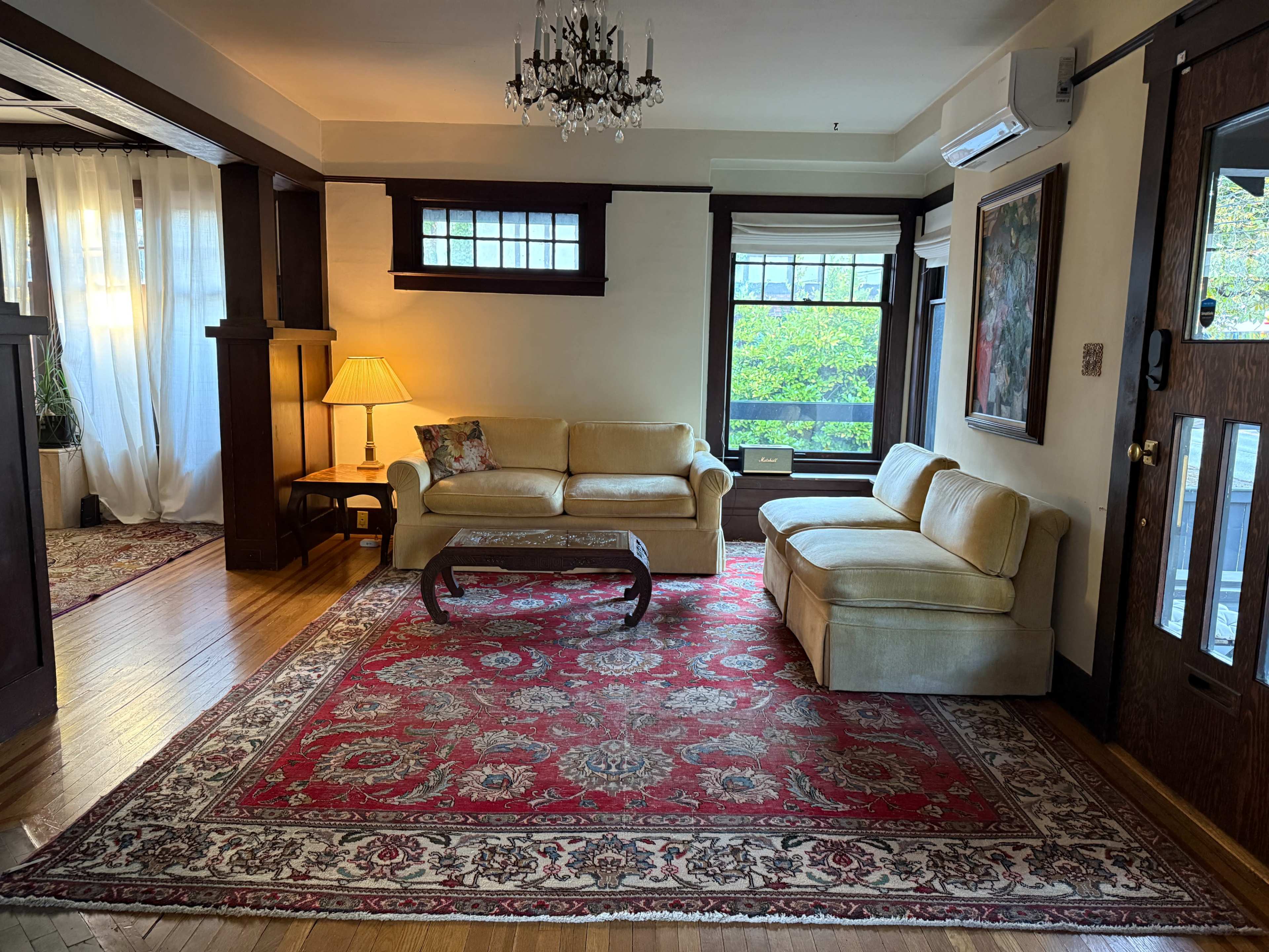 The image shows a living room with a chandelier, a beige couch, a wooden table, a lamp, and a large area rug on hardwood flooring, all illuminated by natural light coming through the windows.